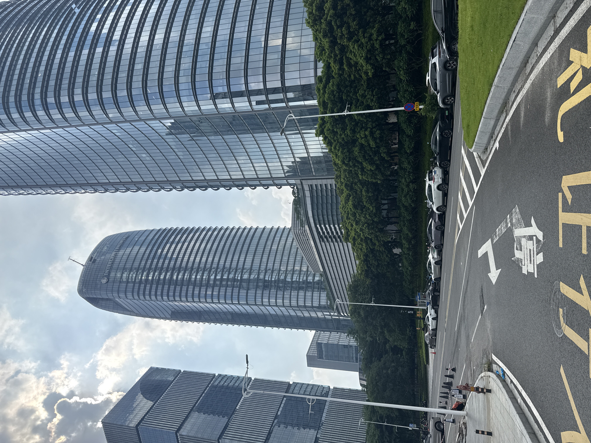 The image shows a street scene in Haizhu District, China, with several modern glass skyscrapers in the background. The buildings are tall and feature reflective facades. In the foreground, a road with lane markings runs through the scene. Several cars are parked along the side of the road, and there are also streetlights and trees lining the street. The sky is partly cloudy. The overall impression is of a contemporary urban environment.