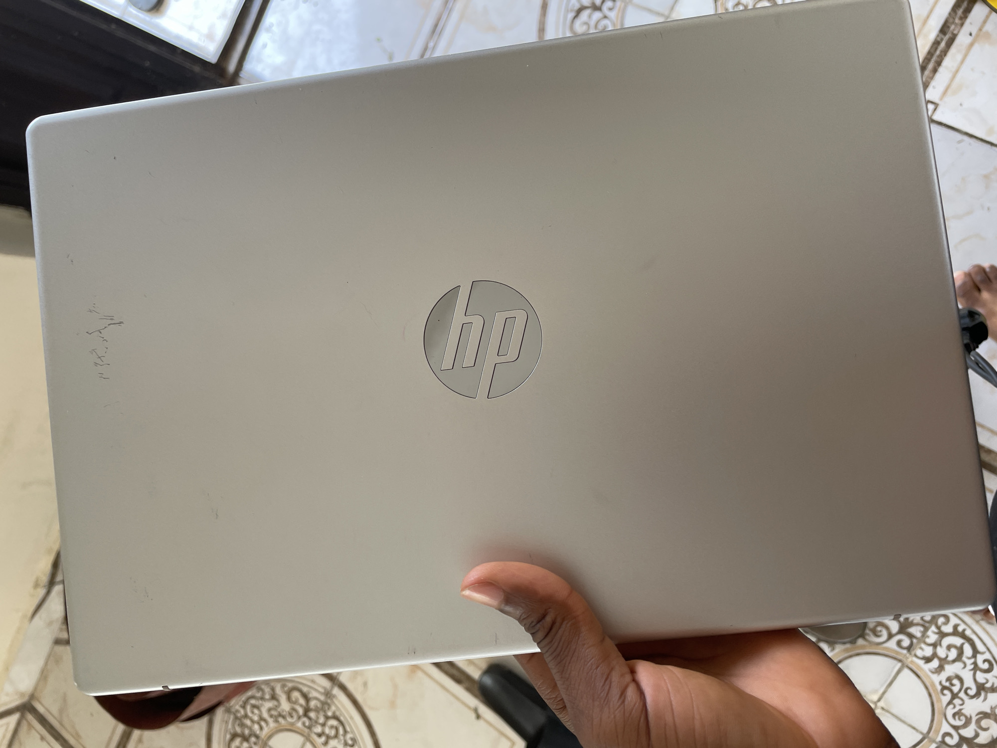 A hand is holding the lid of a silver HP laptop. The HP logo is visible in the center of the lid. The laptop appears to be used, with some minor scuffs and scratches on the surface. The background shows a tiled floor, suggesting an indoor setting, possibly a home or office. A person's hand, with visible fingernails, is supporting the laptop from underneath. The lighting suggests it is daytime. The overall mood is neutral, focusing on the object itself. There is no visible text in the image, other than the HP logo.