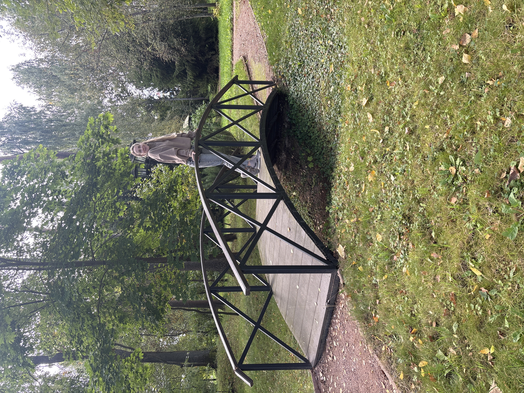 A young woman stands on a wooden bridge with black metal railings in a park in the city of Rozhdestveno, Russia. She is wearing a light brown hoodie and blue jeans, and she is looking at the camera with a slight smile. The bridge spans a small stream or ditch, and is surrounded by trees and grass. The leaves on the trees are green and yellow, suggesting it is autumn. A gravel path is visible to the right of the bridge.