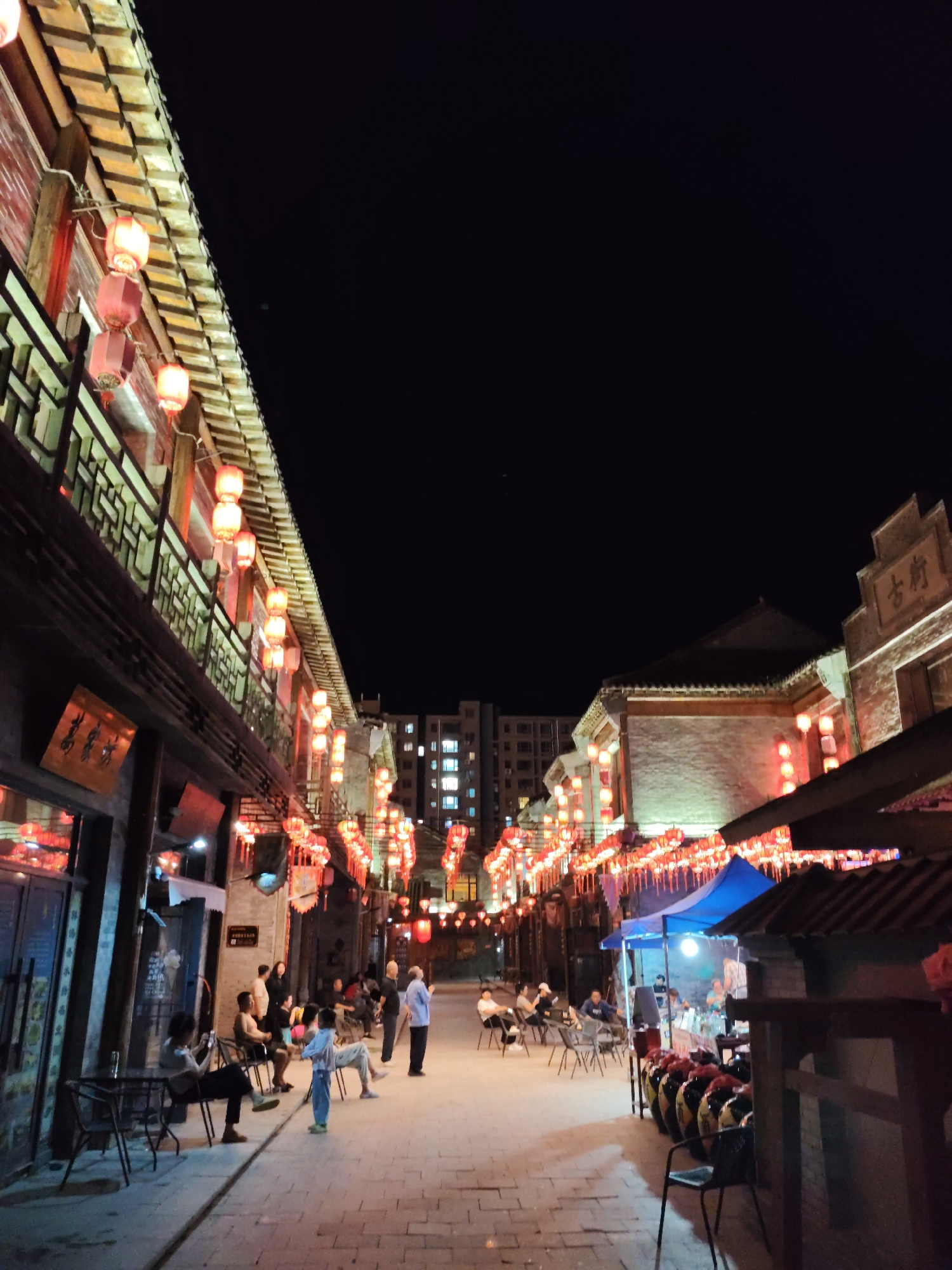 The image depicts a night scene on a pedestrian street in Lindian, China, likely a commercial or historic district. The street is lined with traditional-style buildings adorned with numerous red lanterns, casting a warm glow on the scene. Several people are visible, some seated at tables outside shops or restaurants, while others are standing and walking. In the foreground, a few tables and chairs are set up. Further down the street, tents suggest temporary stalls or food vendors. The overall atmosphere is lively and illuminated, characteristic of an evening in a vibrant urban area. The architecture and lantern decorations point to a cultural or heritage setting.