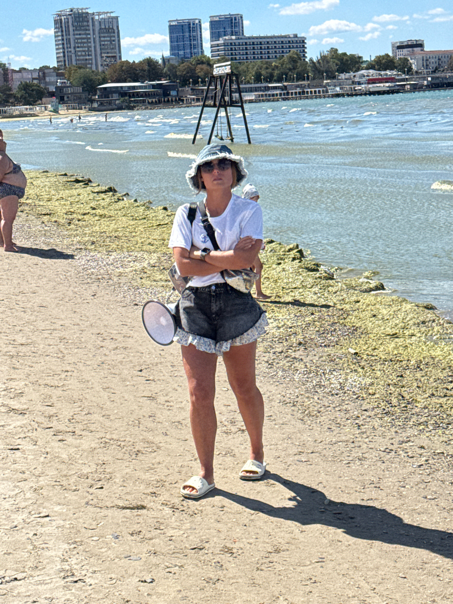 A woman stands on a sandy beach, facing forward. She wears a white t-shirt, dark denim shorts with ruffled edges, a denim bucket hat, sunglasses, and white slides. A gray and silver bag is slung over her left shoulder, and a white megaphone is attached to her right hip. Her arms are crossed in front of her. In the background, the turquoise sea is visible with gentle waves, and a breakwater covered in green algae stretches into the water. In the distance, across the water, are city buildings, including high-rise apartment complexes, and what appears to be a promenade with more buildings along the coast. The sky is blue with scattered white clouds. The scene is set on the beach in Anapa, Russia, on a sunny day.