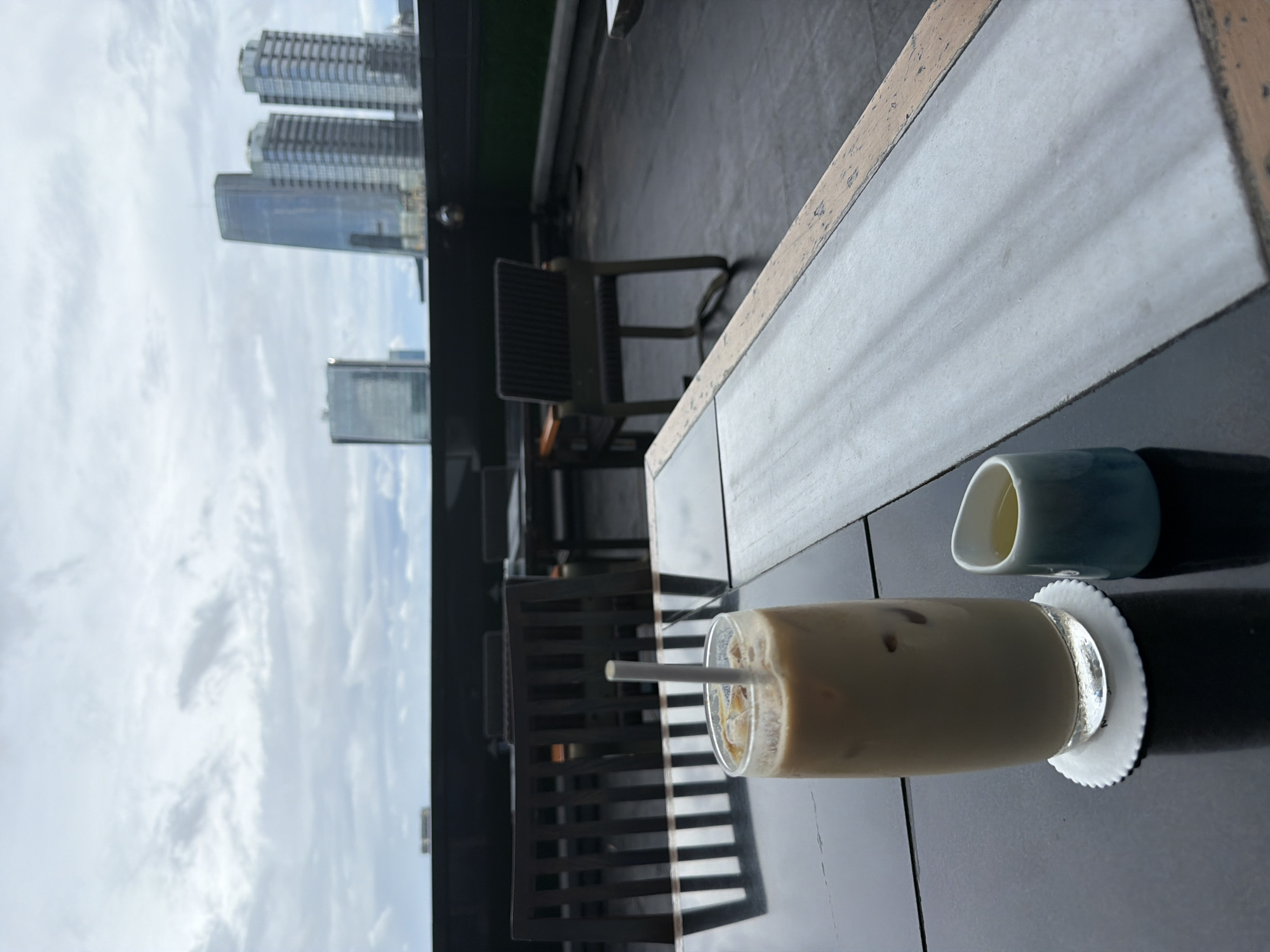 The image shows a rooftop setting, likely in a high-rise building in South Jakarta, Indonesia, as suggested by the distant cityscape visible in the background. The foreground is dominated by a dark-colored table. On the table is a glass of iced coffee with a straw, resting on a white, round coaster. Next to it is a small, blue and white ceramic creamer or sauce dish filled with a light-colored liquid. Chairs are partially visible in the middle ground, suggesting outdoor seating for a cafe or restaurant. The sky is overcast, and the buildings in the distance appear to be modern skyscrapers.
