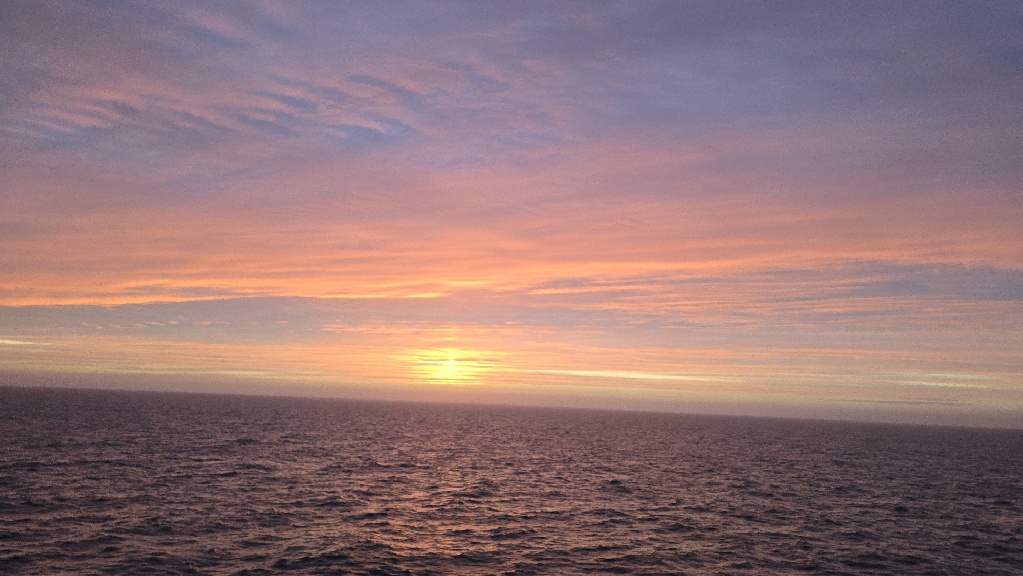 The image depicts a sunset over the ocean. The sky is a gradient of colors, with shades of pink, orange, and purple. The sun is setting on the horizon, creating a bright glow on the water. The ocean is calm with small ripples. The location appears to be offshore in the city of Unknown, Unknown. No people are visible in the image.
