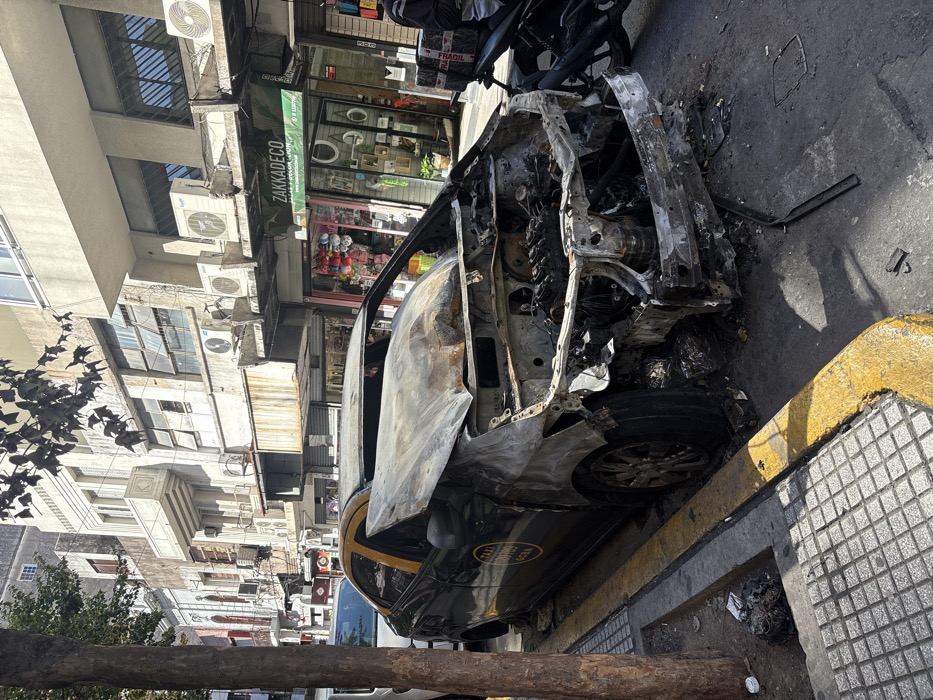 A severely fire-damaged black sedan, identifiable as a taxi by its yellow roof section and partial "TAXI BUENOS AIRES GCBA" decal on the rear door, is parked on an urban street in Buenos Aires, Argentina. The vehicle's entire front section, including the hood, engine compartment, and bumper, is completely destroyed and charred, exposing the engine block and chassis. The interior is also visibly burned, with no glass remaining in the windshield or rear window. The front passenger tire is deflated.
The vehicle rests against a sidewalk featuring gray tactile paving and a yellow-painted curb. Debris, including burnt car components, is scattered on the street and sidewalk around the vehicle. A tree trunk is visible on the far left of the frame, bordering the sidewalk. A white car is partially visible behind the burned taxi, and a black motorcycle is parked directly to its right, with a person's lower body partially visible beside the motorcycle. The motorcycle carries a brown box marked "FRAGIL."
In the background, a row of multi-story buildings lines the street. At ground level, several storefronts are visible, including "ZAKKADECO" with a green sign displaying "www.zakkadeco.com.ar" and another shop featuring various colorful items. The upper floors of the buildings display windows, some with grilles, and numerous external air conditioning units mounted on their facades. The scene is illuminated by daylight.