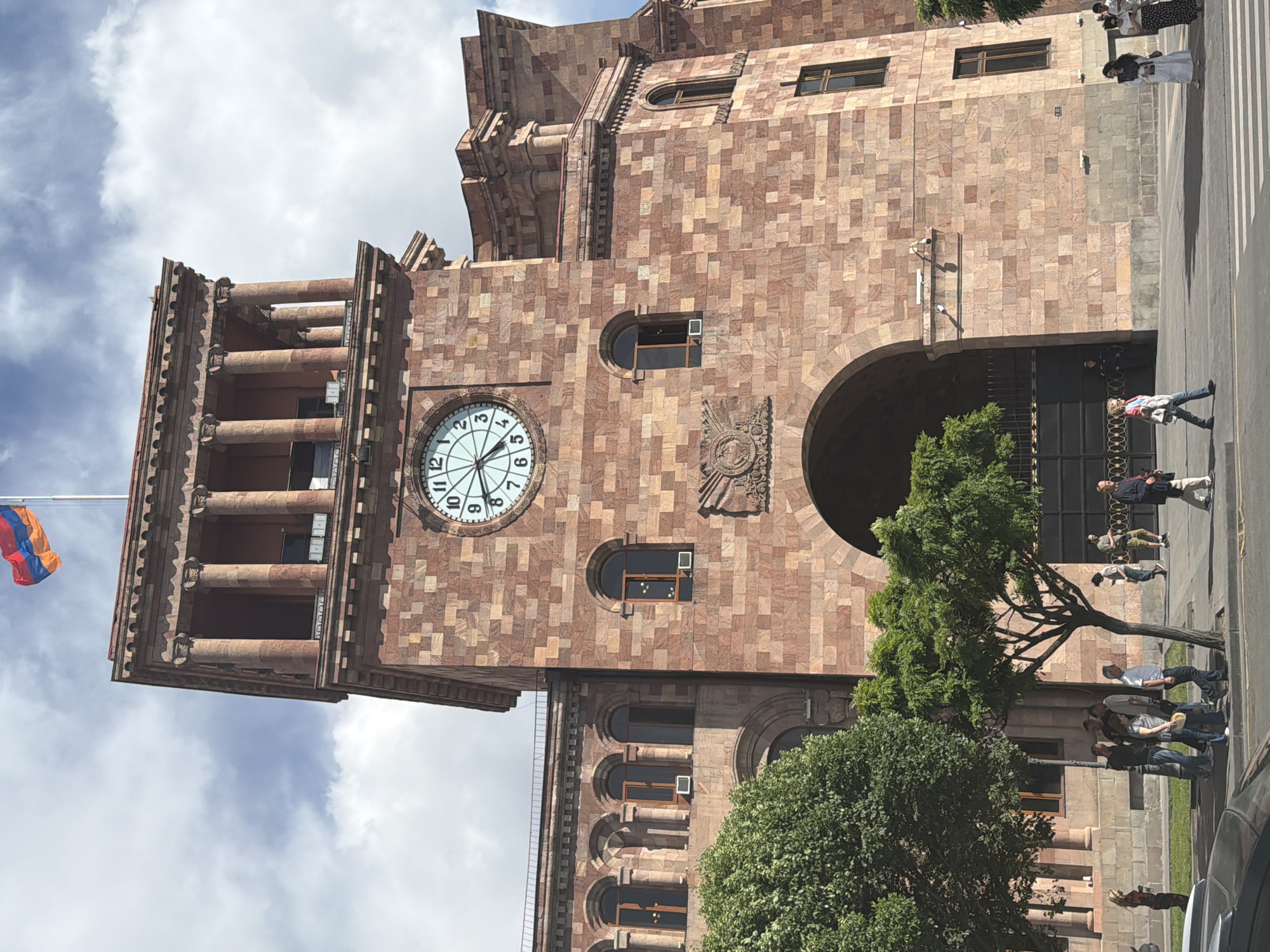 The image depicts the Armenian government building in Yerevan, Armenia, featuring a large clock tower on the left side with a flag of Armenia waving from its top. The building is constructed from red stone blocks and exhibits distinct architectural elements, including columns, arches, and windows. People are observed walking along the sidewalk near the building. Trees are planted near the building.