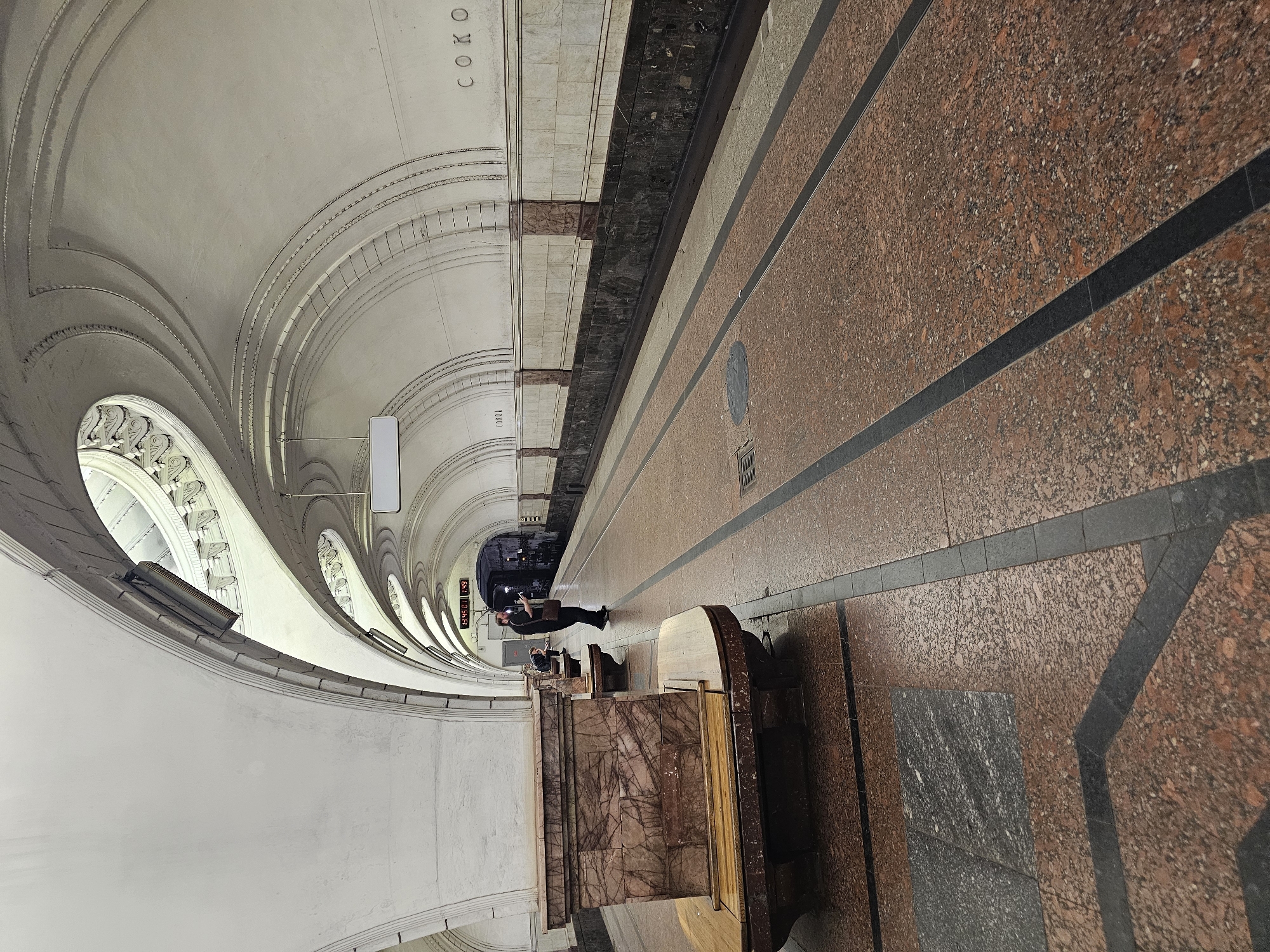 This is a medium-angle, eye-level shot of a metro station platform in Moscow, Russia. The platform is lined with benches made of marble and wood, and a man in a dark shirt and pants stands on the platform, looking at his phone. The station has a distinctive architectural style with a curved white ceiling and arched passages. The floor is paved with red and grey granite tiles. In the distance, a train is visible at the platform. The station is named "Sokolniki".