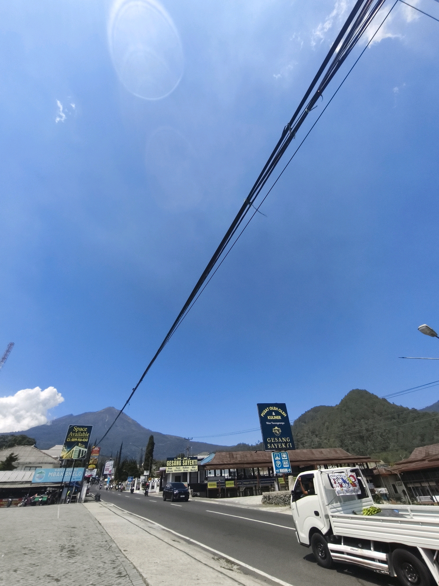 The image depicts a street scene in Tawangmangu, Indonesia, with a large mountain dominating the background. The sky is a clear, bright blue with a few wispy clouds and a noticeable lens flare or light reflection in the upper left quadrant. Overhead, a tangle of dark power lines stretches diagonally across the frame.
In the foreground, a paved road runs from left to right, with a concrete sidewalk bordering its left side. Several vehicles are present: a black SUV is driving on the road, and a white flatbed truck with a person visible in the driver's seat is also on the road, its bed containing what appears to be produce. Parked vehicles, including motorcycles, are visible on the sidewalk to the left.
Along the roadside, there are various buildings and commercial establishments. Prominent signs are visible, including one that reads "Space Available CS: 0899-906-4243". Another large blue sign states "PUSAT OLEH OLEH & KULINER Tawangmangu" which translates to "Souvenir and Culinary Center Tawangmangu", and below it "GESANG SAYEKTI". There are also smaller banners and signage for shops and businesses.
The architecture of the buildings appears to be a mix of traditional and modern styles, with some structures featuring wooden elements and tiled roofs. The overall impression is of a commercial area situated in a mountainous region, likely a tourist destination given the "Souvenir and Culinary Center" sign. The presence of the mountain, road, buildings, and vehicles suggests a populated, active environment.
