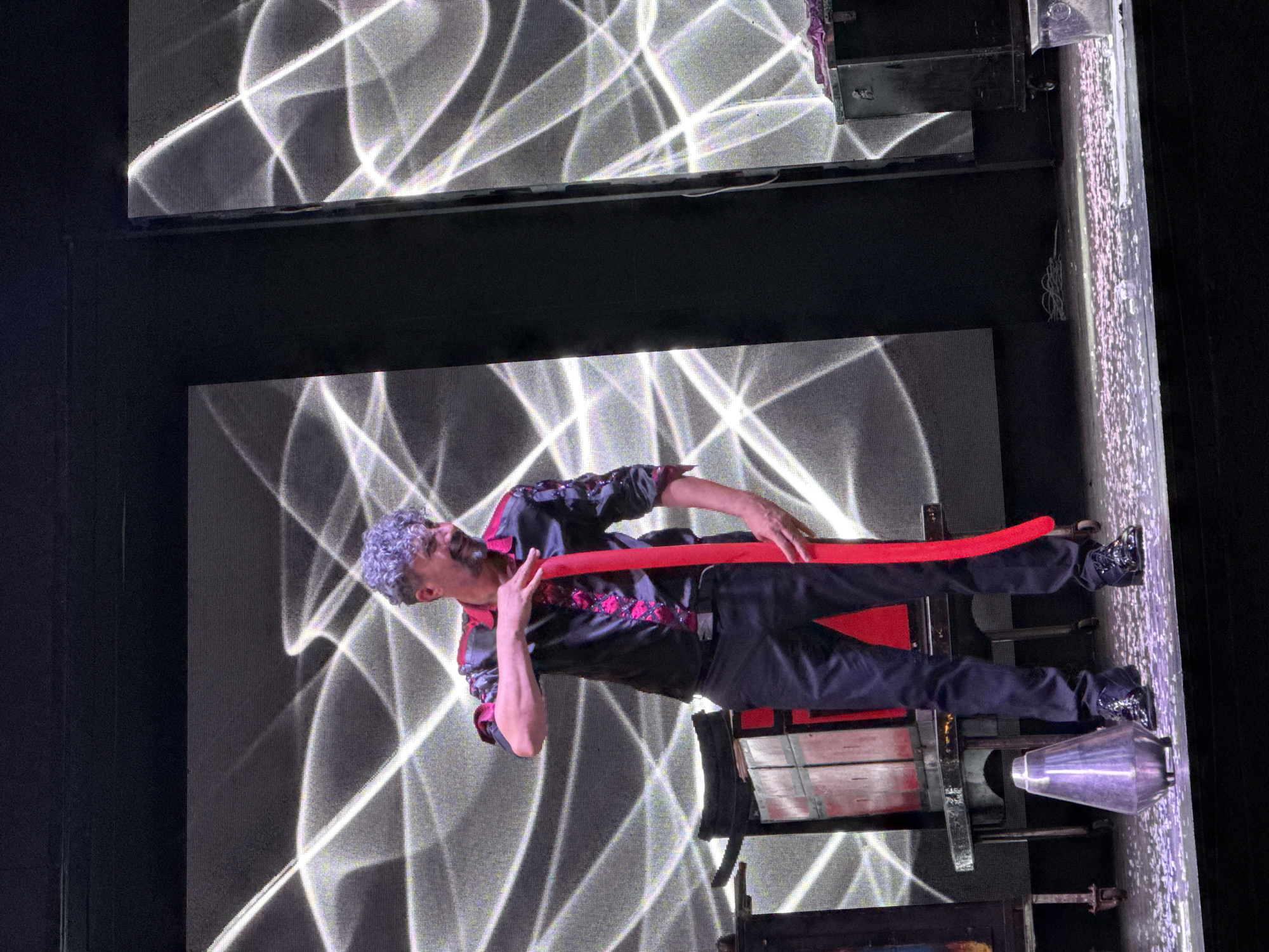 A magician with curly gray hair and a beard, wearing a black and pink patterned shirt and black pants, stands on a stage and holds a long, red balloon. He is looking down and to his right with a slight smile. Behind him, a large screen displays a dynamic white light pattern against a black background. To his left, a stage prop resembling a red and black box is visible. The stage floor is covered with confetti. A metallic, cone-shaped object rests on the stage near the magician's feet. The overall environment appears to be a performance venue, possibly a theater or entertainment hall. There is no information about the location being in Manavgat, Turkey.