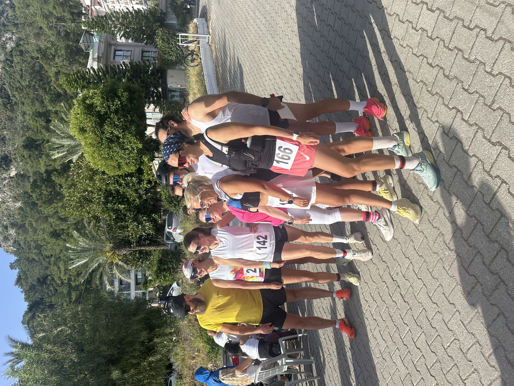A medium shot shows nine people standing side by side on a gray-paved area. The people are all dressed in athletic wear and appear to be getting ready for a race, as some are wearing bib numbers. Starting from the left, the first person is wearing a yellow t-shirt and black shorts with red running shoes. The next person is wearing a white t-shirt and black shorts with the bib number 2. Then there is another person in a white t-shirt and black shorts with bib number 142. The next person is wearing a white t-shirt and pink sports bra with bib number 166. Finally, there is another person in a white tank top and black shorts. The background of the image is blurred, but it appears to be a residential area with trees and buildings. The location may be in the city of Fethiye, Turkey.