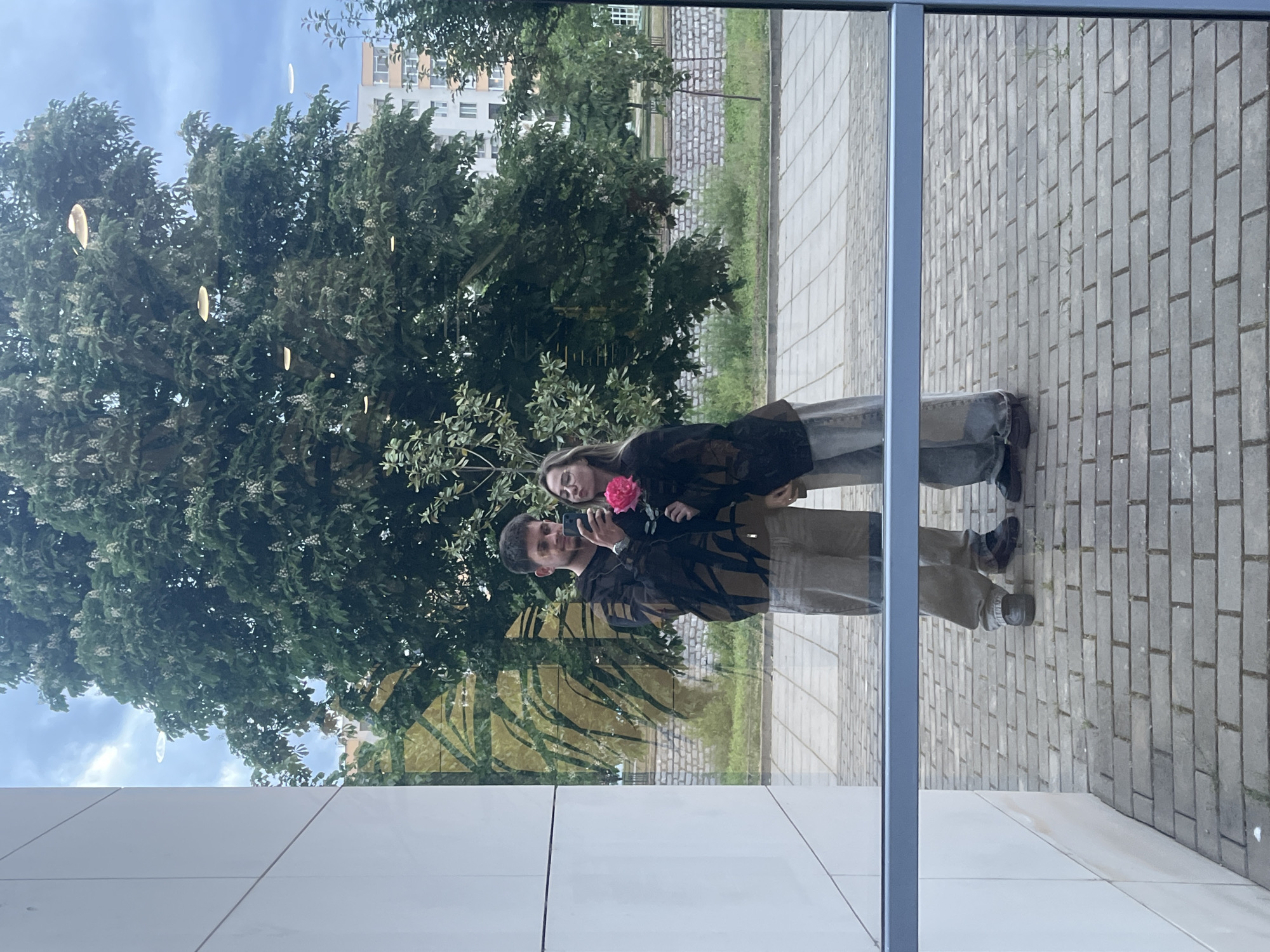 The image depicts a couple standing in front of a mirrored glass wall, likely a building facade. Their reflections are visible in the glass, showing a man in a brown jacket holding a phone, possibly taking a picture, and a woman with a pink flower. The background reflection shows a tree, a building, and a paved area, suggesting an outdoor location. Based on the urban architecture and vegetation in the reflection, the location may be Blagoevgrad, Bulgaria. The primary subject of the photo is the reflection of the couple in the mirrored glass.