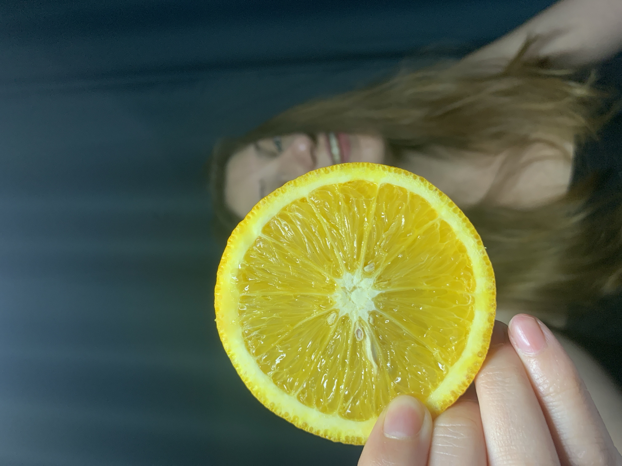 A close-up shot features a slice of orange held up against a dark, blurred background. A hand with pink nail polish holds the orange slice. Behind the orange, the blurred image of a woman with blonde hair and a smile is visible. The orange slice is in sharp focus, highlighting its juicy texture and radial segments. The background appears to be dark and possibly textured. There is no information in the image that indicates this was taken in Chaikovsky, Russia.
