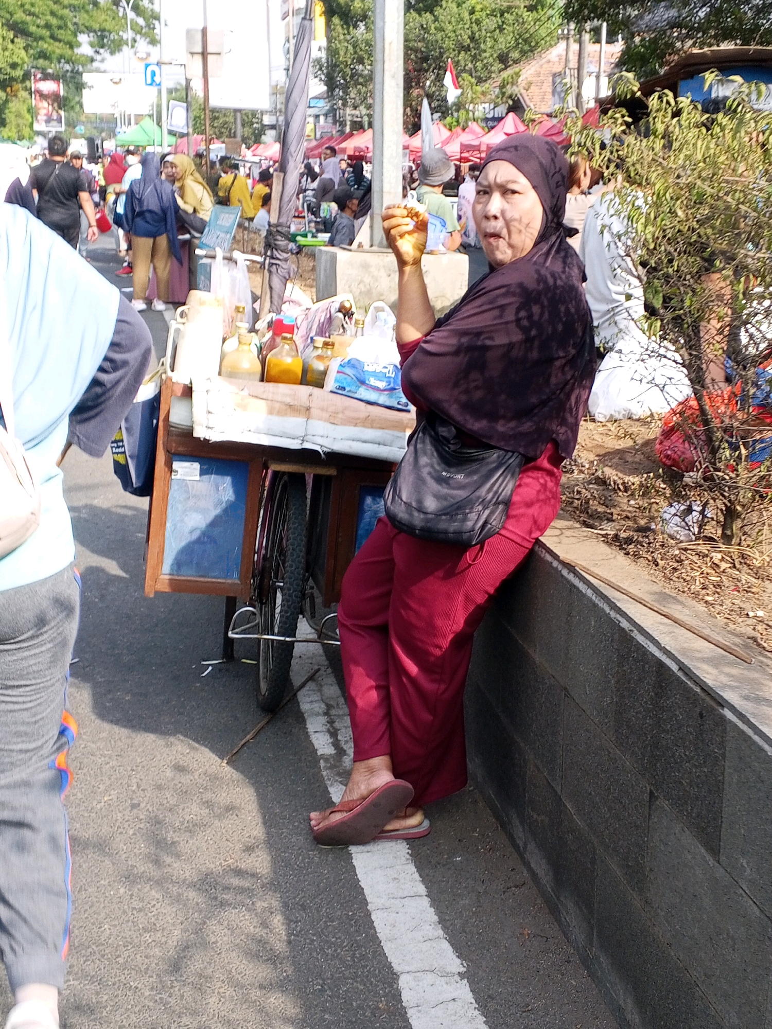 The image depicts a street scene in Serang, Indonesia, likely a market or roadside area. In the foreground, a woman wearing a dark headscarf and maroon pants is leaning against a low stone wall. She is holding a small object in her right hand, which appears to be a piece of food or a small trinket. Her left arm is resting on her knee, and she has a black crossbody bag slung over her shoulder. To her left, a portion of another person is visible, wearing a light blue long-sleeved shirt and gray pants.
Behind the woman, a cart is positioned on the side of the road. The cart has a wooden frame and is laden with various items, including bottles filled with liquids, a white pitcher, and possibly some packaged goods. The cart's wheels are visible, one of which is partially obscured by the woman's body. The cart appears to be a mobile vendor's stall.
In the background, a bustling scene unfolds with several other people and market stalls. Tents and awnings are set up, suggesting an outdoor market environment. People are seen walking and interacting. The area is characterized by roads, some vegetation, and buildings in the distance. The overall atmosphere appears to be that of a busy public space during daylight hours.