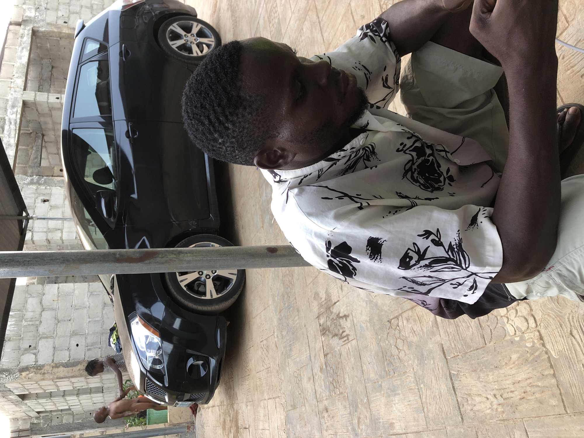 The image shows a black car parked on a patterned concrete surface. A man in a patterned white and black shirt and khaki shorts is seated to the side of the car. Two figures, possibly children, are seen near the front of the car. The setting appears to be outdoors, possibly in a residential area, evidenced by the block wall visible in the background. The city and specific location are Unknown.