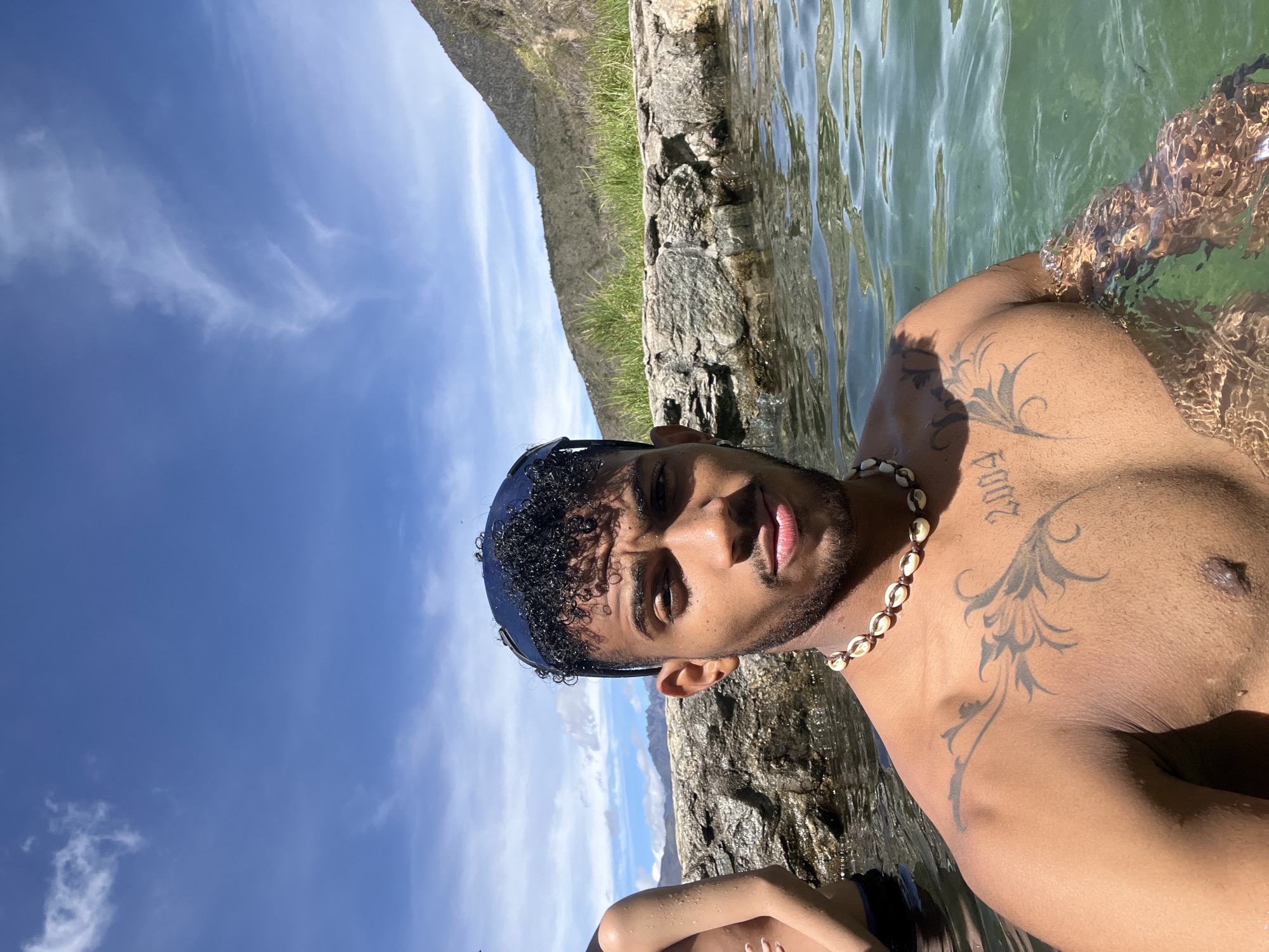 A medium-shot, front-facing selfie shows a young man with dark, curly hair and a beard, wearing sunglasses pushed back on his head and a seashell necklace. He is submerged in clear, green water, with a rocky wall and vegetation in the background, under a bright blue sky with scattered clouds. A portion of another person's torso is visible to the left. The man has tattoos on his chest and left shoulder. The scene appears to be an outdoor natural setting, possibly a hot spring or a natural pool, consistent with the geography of Mucurubá, Venezuela, which is known for its mountainous terrain and natural springs.