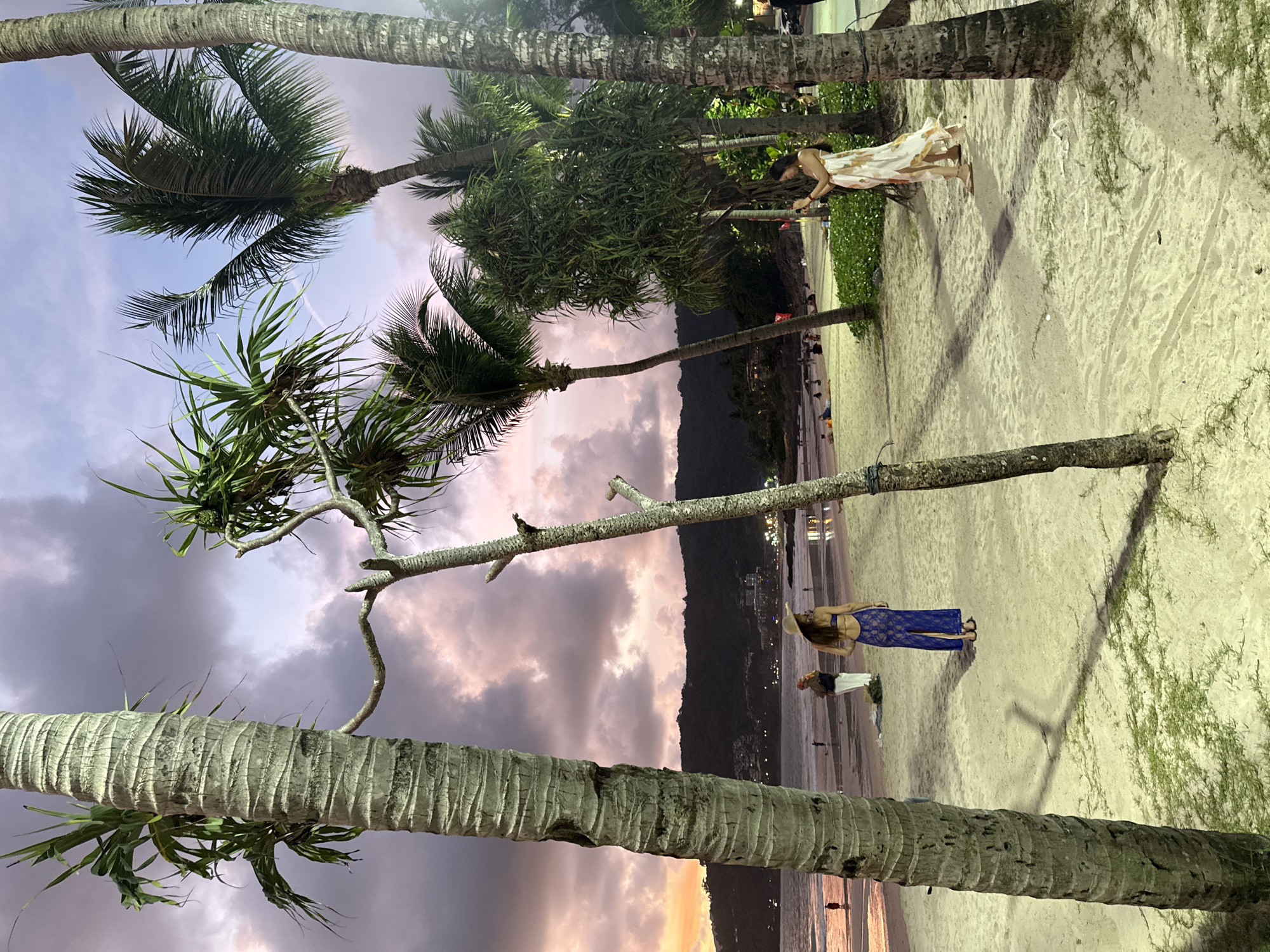 At dusk on a beach in Patong, Thailand, two women are visible. The first woman, wearing a blue lace dress, stands facing away from the camera, her back to the ocean. The second woman, to her right, is captured from behind and side, holding a cell phone and taking a picture of the sunset. She is wearing a light-colored, patterned maxi dress. Palm trees frame the scene, with the sand stretching towards the ocean where faint figures can be seen in the water. The sky is a gradient of purple and pink, with scattered clouds. Lush green foliage lines the edge of the beach, bordering a walkway.