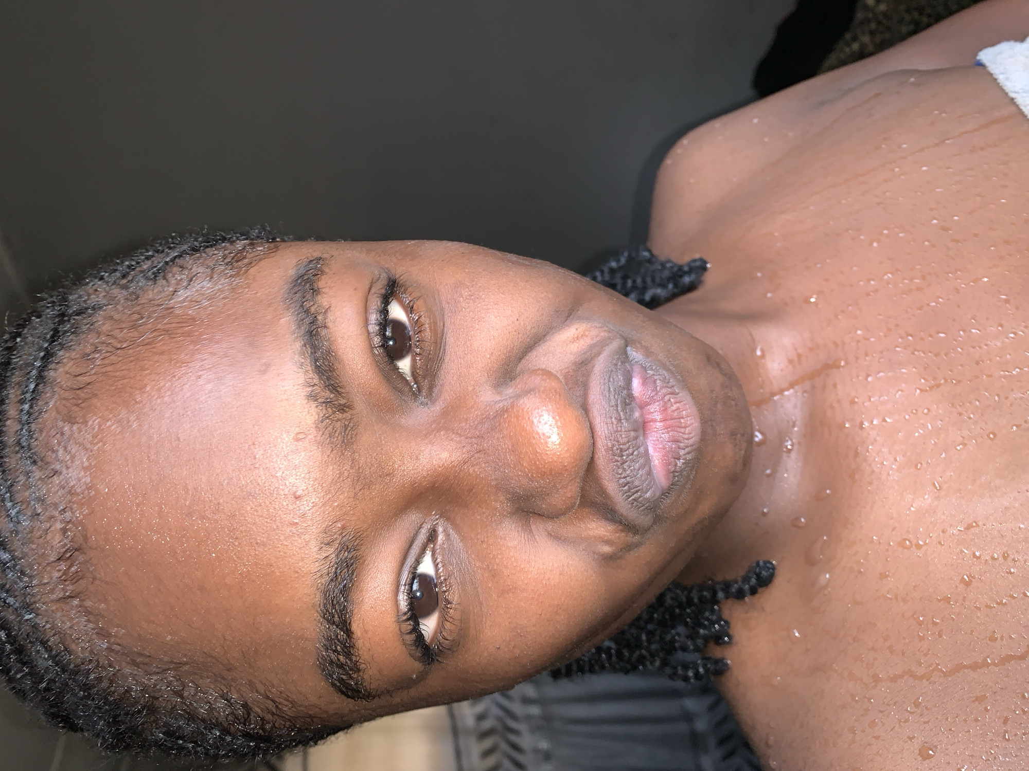 A close-up portrait, rotated 90 degrees clockwise, features a person with dark skin, likely female, captured from the chest up. The individual's face is prominent, with dark brown eyes gazing directly at the camera, framed by long, dark eyelashes and well-defined eyebrows. The lips are slightly pursed, displaying a textured, pinkish hue. Dark, tightly braided hair frames the face and extends onto the shoulders.
A notable detail is the presence of numerous small water droplets covering the person's forehead, cheeks, shoulders, and bare upper chest, suggesting they have recently bathed, are sweating, or are in a humid environment. A small portion of a white towel or cloth is visible near the right shoulder, reinforcing the idea of a post-bath moment. The background is dark and indistinct, indicating an indoor setting with low or artificial lighting, which creates highlights on the wet skin. The expression is neutral, possibly thoughtful or slightly playful due to the pursed lips. The scene is likely set in Rafin Par, Nigeria. No text is visible in the media.