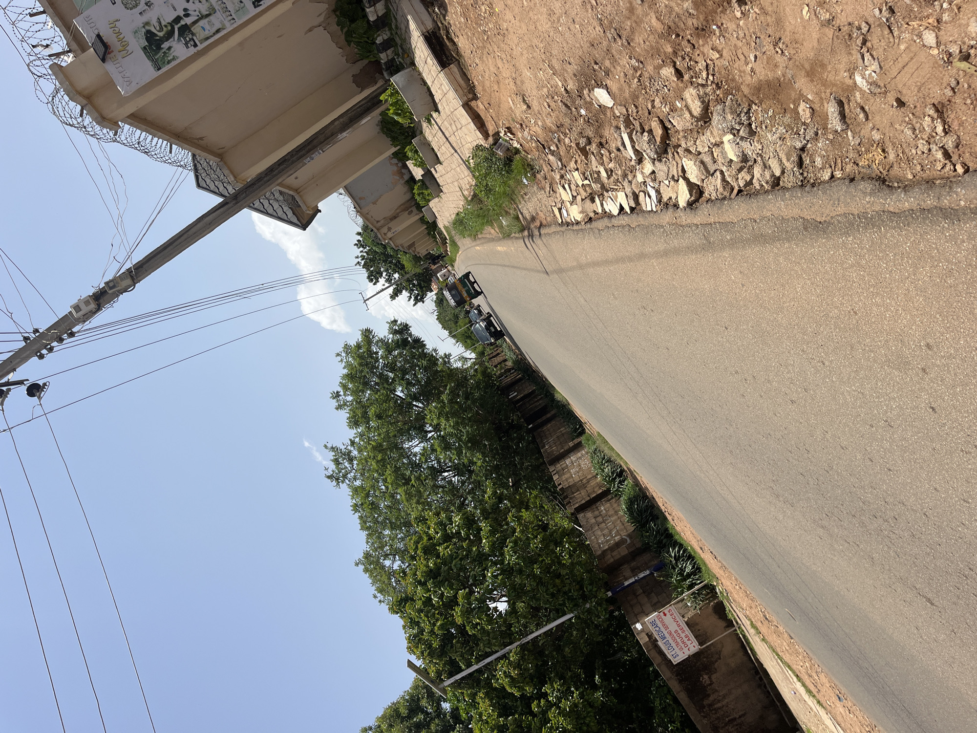 The image shows a street in Gura Top, Nigeria. The street is paved and appears to be a residential or commercial area. The sky is blue with a few scattered clouds. There are trees and buildings on either side of the street, and there are power lines running overhead. A few vehicles, including what appears to be a tuk-tuk and a car, are visible on the road. There is a sign for "St. Louis Medicare" with services listed as "Ultrasound Services, Drugs, Lab Services". The buildings are modern with security features such as barbed wire on the walls. The ground is a mix of asphalt and dirt, with some rocky debris along the roadside.
