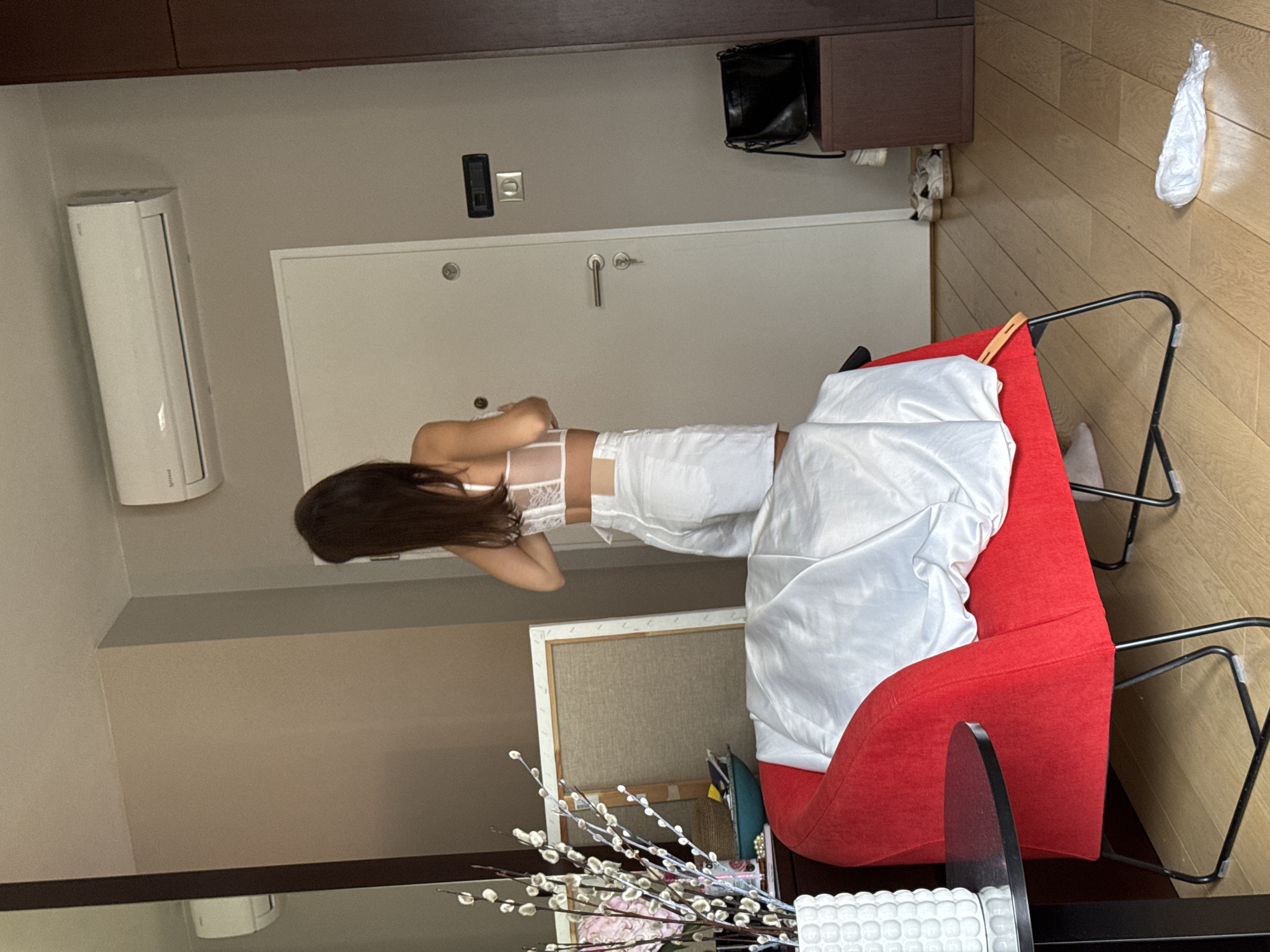 A person lies on a red chair in a room. The person has long, dark hair and wears white lingerie. A white blanket is draped over the chair. In the background, there is a door, an air conditioner, and wooden furniture. A plastic bag hangs on the wooden wall. There are flowers in the foreground. The location is Unknown, Unknown.