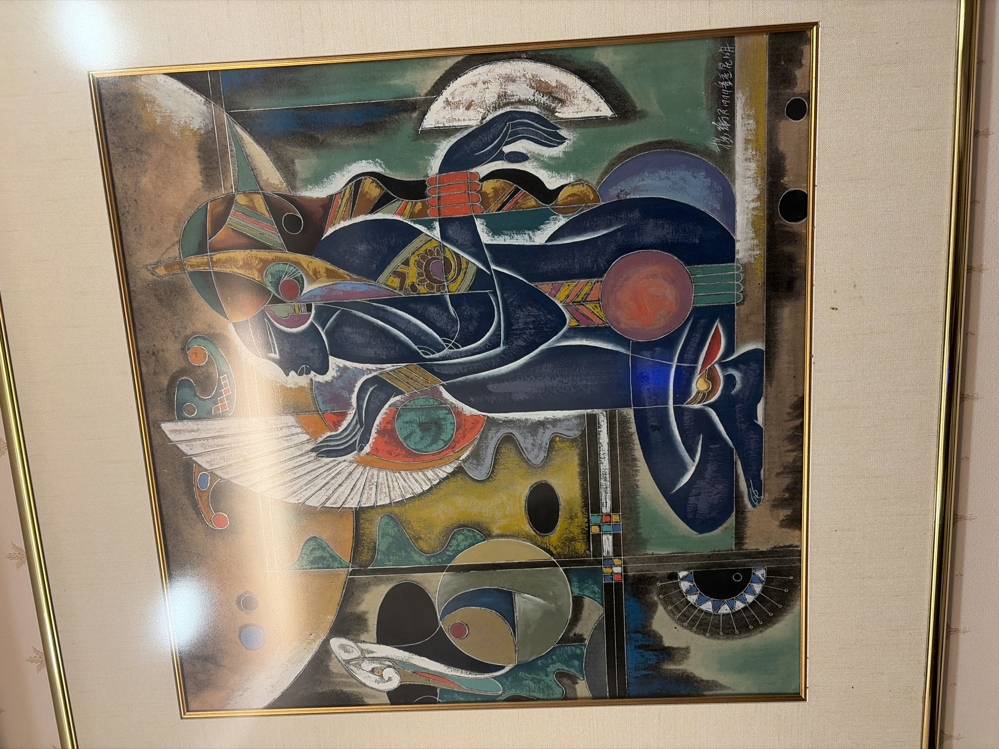 The image is a framed painting depicting an abstract representation of two figures in profile, rendered in dark blue with colorful accents. The figures are stylized and appear to be seated or kneeling, facing left. The left figure's head is tilted back, and its hands are raised. The right figure is behind and to the right of the first, with its head also in profile and one arm extended.
The background of the painting is a mosaic of geometric shapes and organic forms in muted tones of brown, yellow, green, and blue, with some areas of stark white and black. There are elements that suggest fans or wings, as well as circular motifs and angular lines. The overall style is abstract and somewhat cubist, with a focus on color and form.
The painting is framed in a thin gold metal frame, and it is matted with a textured beige material. The artwork itself appears to be acrylic or oil on canvas or board. There is a signature and date in the bottom right corner of the painting, which appears to read "楊衡民 1994" and some other characters. The image of the painting is taken in a horizontal orientation, and it is lit by overhead lighting, which causes a reflection on the upper left portion of the frame and the artwork. The painting is hanging on a wall with a subtle patterned wallpaper. The setting is indoors and appears to be a domestic or gallery environment, though the specific city and location are unknown.