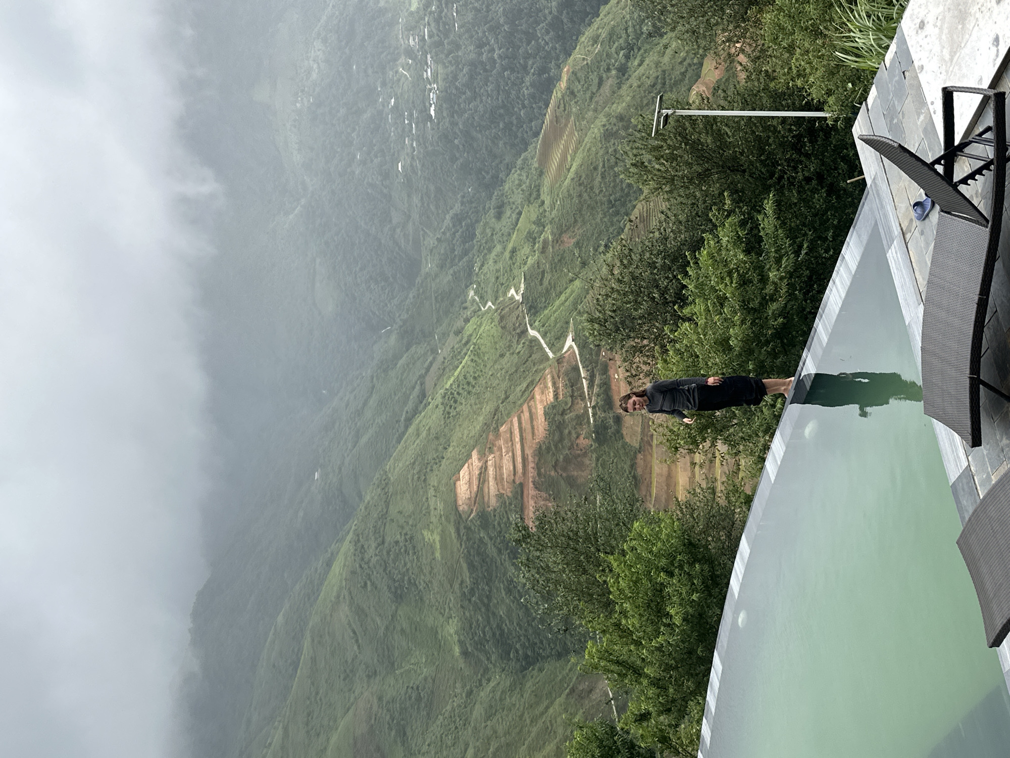 A lone woman stands at the edge of an infinity pool, her reflection visible in the calm green water. She is wearing a dark long-sleeved top and black skirt, with her feet bare. The pool overlooks a vast, verdant mountain landscape in Ta Xua, Vietnam, shrouded in mist. Terraced fields and a winding road are visible on the mountainside. In the foreground, two dark lounge chairs are positioned on a stone patio next to the pool. A single blue flip-flop rests on one of the chairs. The scene conveys a sense of tranquility and awe amidst the natural beauty of the mountainous region.
