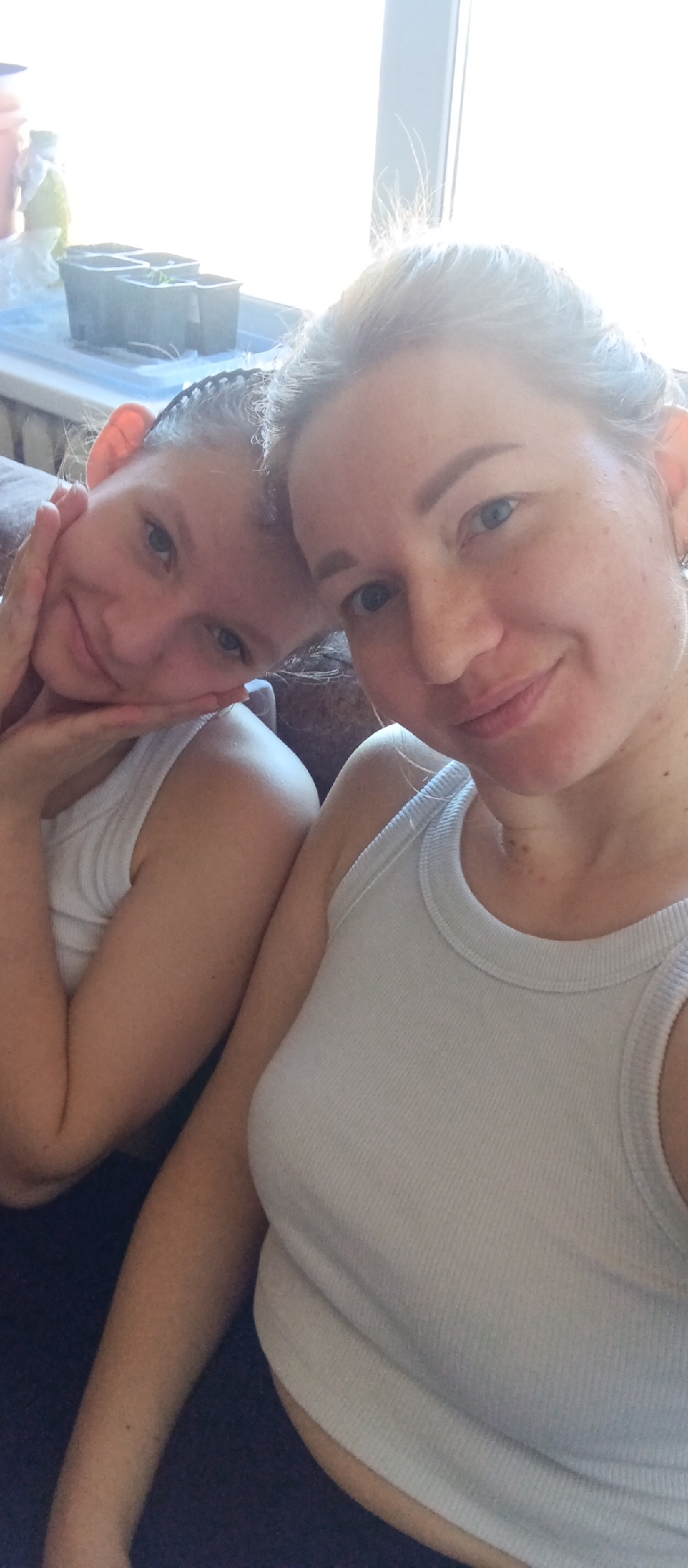 Two females, a woman and a girl, are in a well-lit indoor environment. Both are wearing white tank tops. The girl is resting her head on the woman's shoulder, her hand gently touching her face. There are small potted plants visible in the background. The location appears to be in the city of Unknown, Unknown.