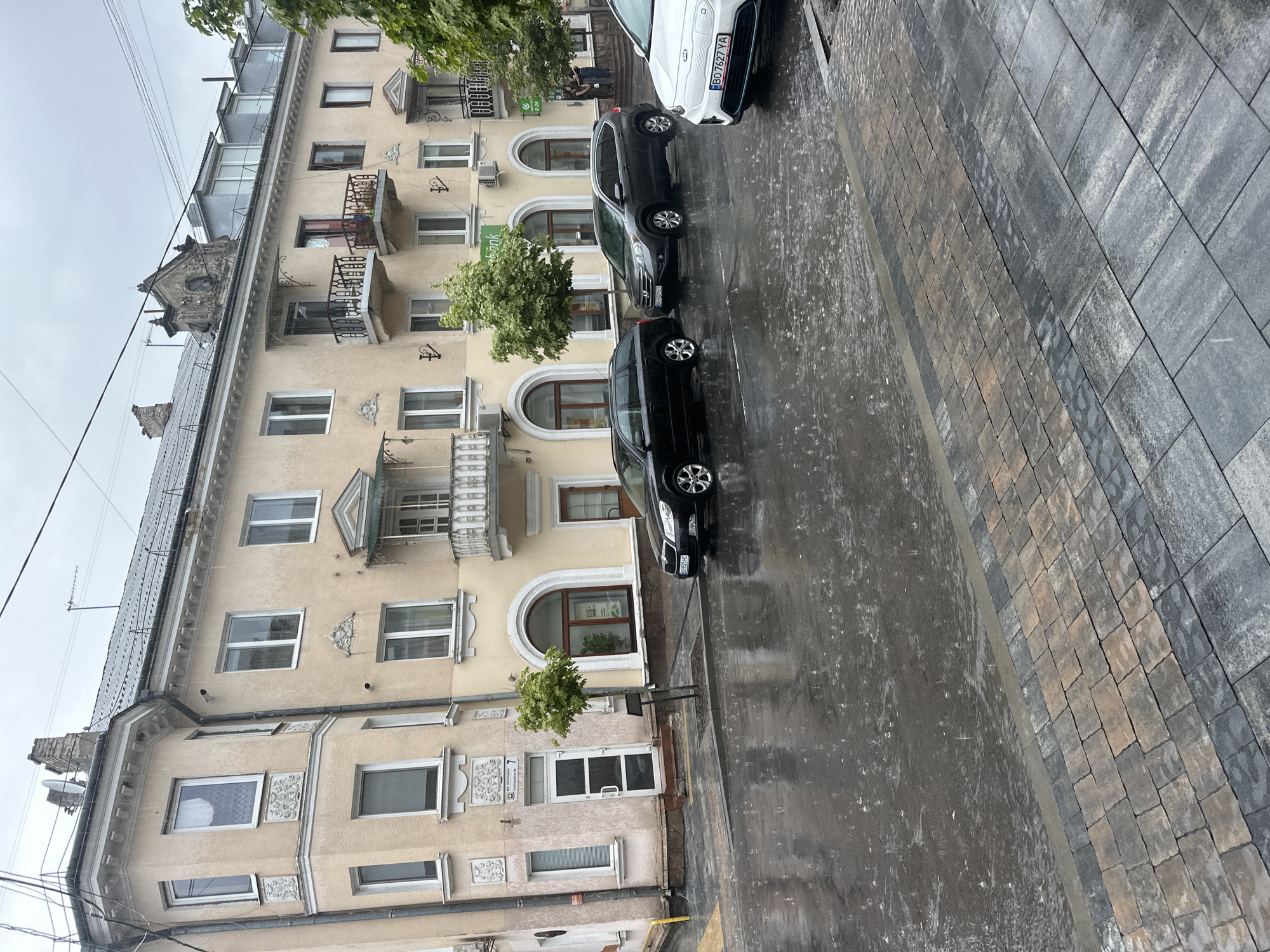 This image depicts a street scene in Ternopil, Ukraine, during a rain shower. The primary focus is a pale yellow, multi-story building with ornate architectural details, including balconies with wrought-iron railings and decorative window surrounds. Several windows are visible, some with curtains. A few trees, one of which is small and green, are planted along the sidewalk.
The street in front of the building is wet and reflects the overcast sky and surrounding structures, with visible raindrops creating ripples on the surface. The sidewalk is paved with interlocking bricks in various shades of grey and brown.
Parked on the street are three vehicles. From left to right, these are a black station wagon, a dark gray SUV, and a white electric vehicle with a license plate that reads "BO 7627 YA". The white vehicle appears to be a Kia.
In the background, on the right side of the frame, a few people are standing near the entrance of a building, possibly taking shelter from the rain. The sky is overcast, suggesting a cloudy or rainy day. Electrical wires are strung across the top of the image.