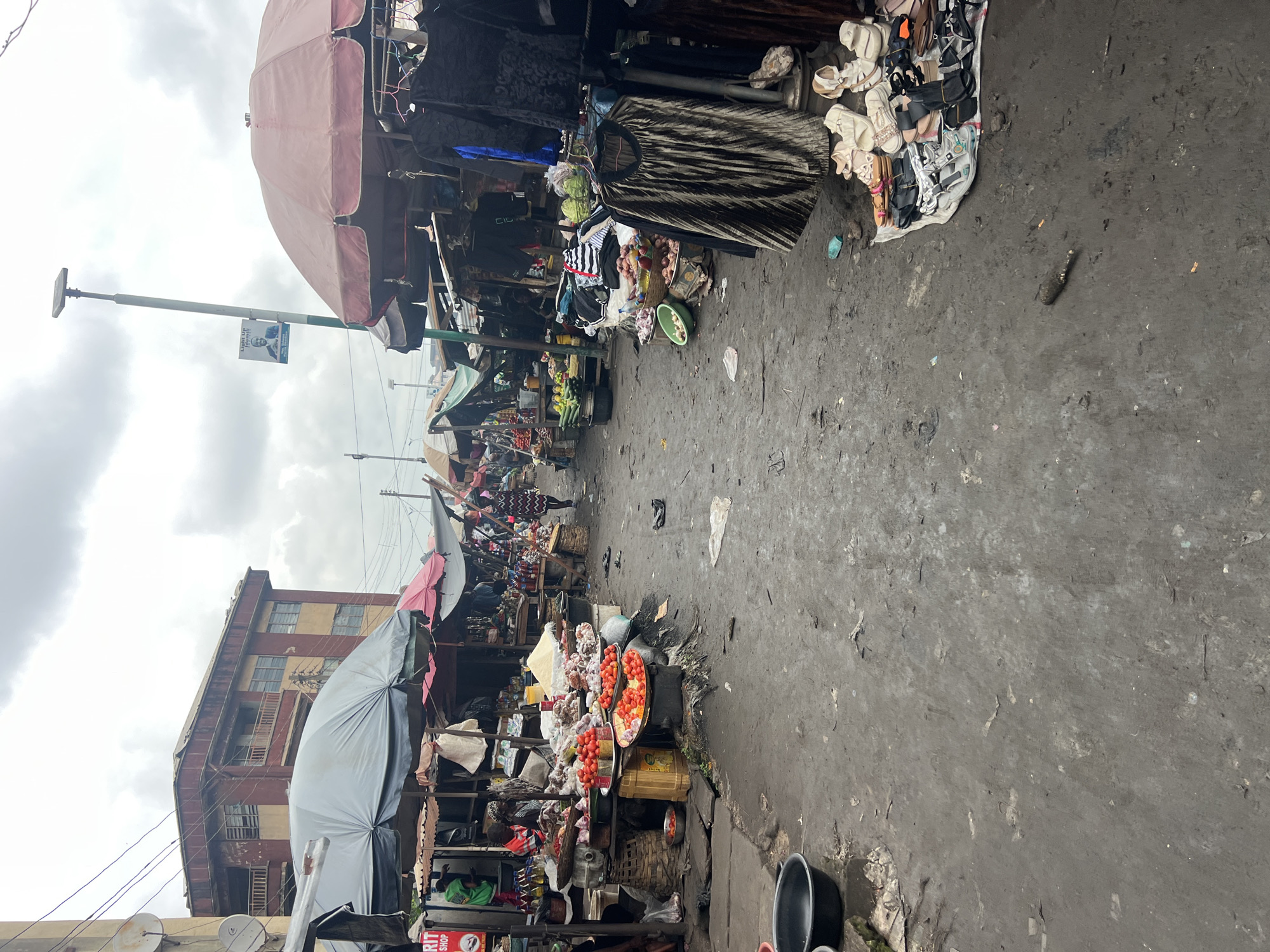 The image depicts a bustling outdoor market in Nkpor, Nigeria, under an overcast sky. The market is lined with stalls selling various goods, including produce, clothing, and shoes. The ground is muddy and littered with debris, suggesting recent rain or a lack of regular cleaning. In the foreground, a large black basin sits on the left, partially filled with water. A woman in a patterned dress walks away from the viewer down the center of the market. Other individuals are visible in the background, engaged in market activities. The stalls are covered with tarps and umbrellas for shade, with clothing racks and tables displaying goods. A lamppost with a political poster is visible in the distance. The overall impression is one of a vibrant, active marketplace, despite the somewhat unkempt environment.