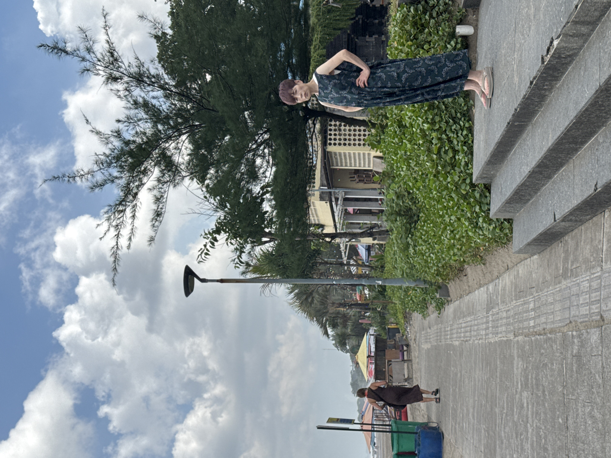 This outdoor image, taken in Kuta, Indonesia, captures a bright, partly cloudy day. The scene features a paved walkway bordered by lush green foliage and various trees, including tall, slender-needled varieties and palm trees.
In the mid-ground on the right side, a woman with short, reddish-brown hair is standing on a concrete platform, partially elevated by a few steps. She is wearing a dark green sleeveless dress with a light pattern and light-colored sandals. She poses for the camera with one hand on her hip, looking directly ahead.
Further down the walkway, towards the foreground on the left, another person in dark clothing is seen from behind, pulling two wheeled containers, one blue and one green. The pathway itself has textured sections, indicating a designated pedestrian area. Along the side of the path, obscured by vegetation in places, are various small structures or stalls, some with visible colorful awnings. A street lamp post stands on the left. The sky is clear blue with scattered white cumulus clouds. No legible text is visible in the image.