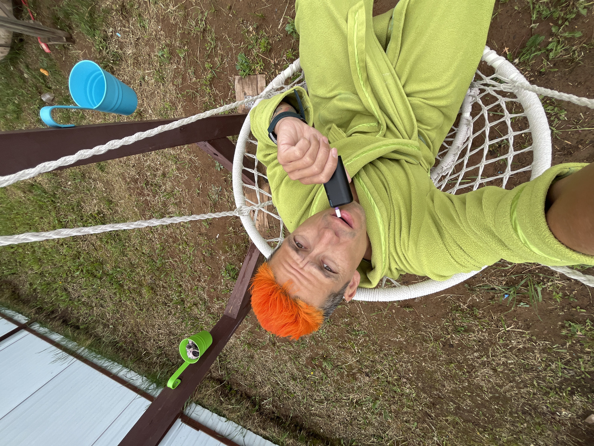 A man lies in a white rope hammock in a grassy, slightly muddy yard. He is wearing a green robe and has bright orange hair on top of his head. He is holding a black electronic device to his lips. A dark brown wooden frame supports the hammock. Blue and green plastic cups sit on the frame. Part of a white fence is visible along the bottom edge of the image. The location is Chaikovsky, Russia.