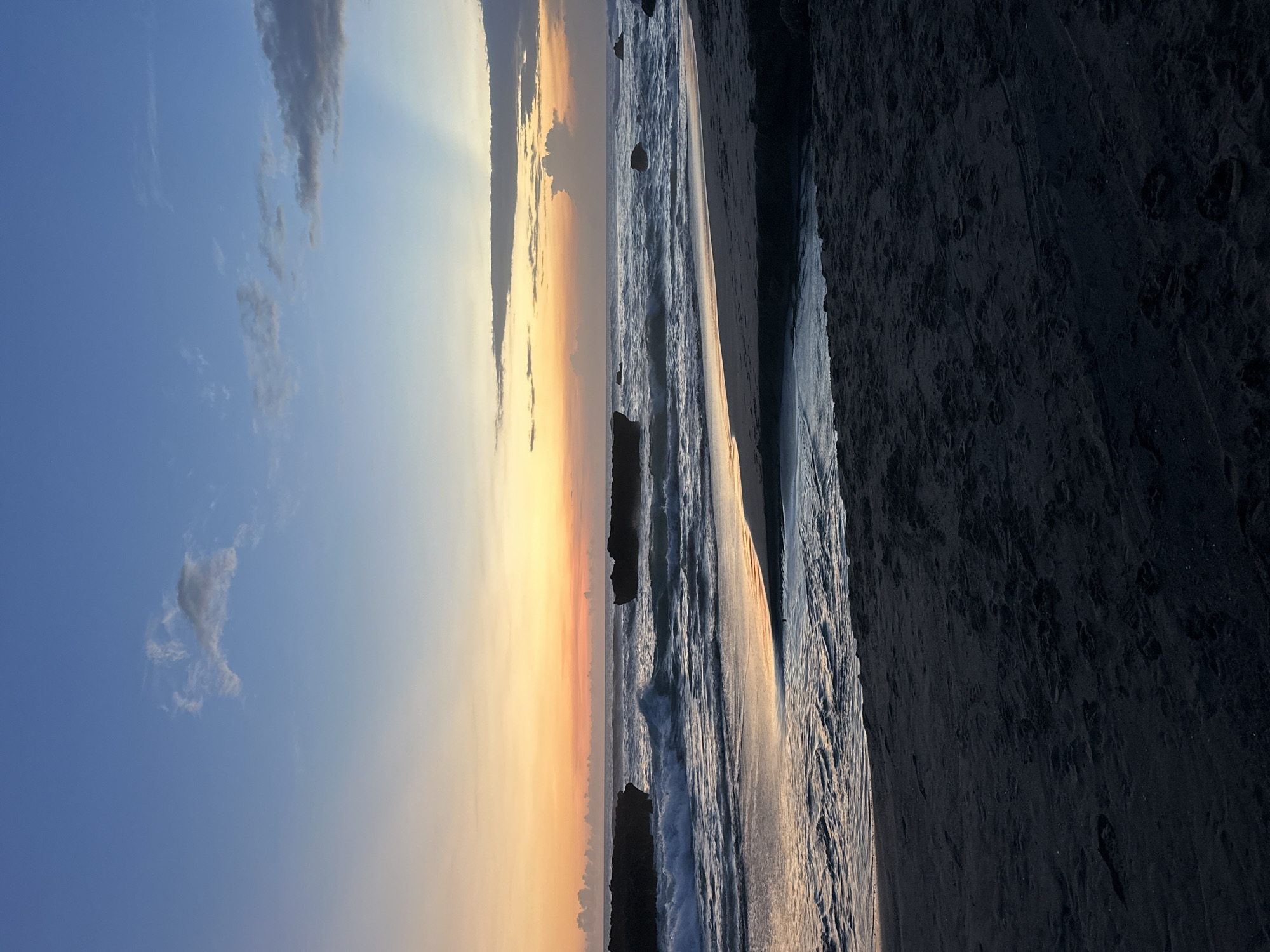 The image depicts a sunset scene at a beach in North Kuta, Indonesia. The sky transitions from blue with scattered clouds to orange and yellow hues near the horizon. The sea is visible with gentle waves approaching the shore. Several dark rock formations are present in the water near the shoreline. A dark sandy beach stretches along the right side of the frame. No people or animals are visible.