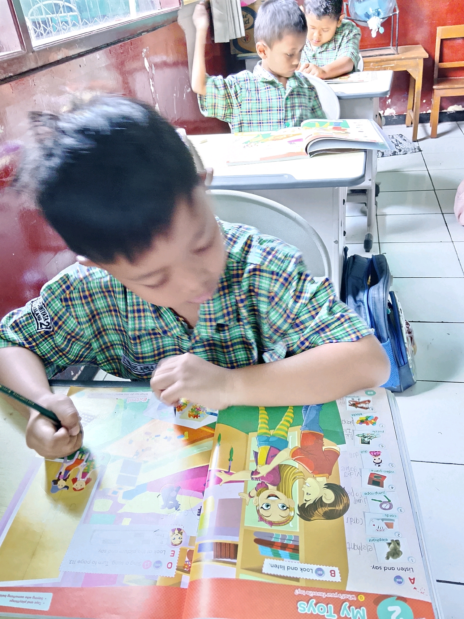 In Malang, Indonesia, two young boys in plaid shirts sit at desks in a classroom. The boy in the foreground is leaning over a book, with his left hand on the book and his right hand holding a pencil, appearing to be drawing or writing. He has dark hair and is wearing a green, blue, and yellow plaid shirt. A blue backpack is visible behind him, leaning against his chair. The boy in the background, also wearing a plaid shirt, is looking down at his book. The classroom appears to have windows with patterned glass and wooden furniture. The floor is tiled.