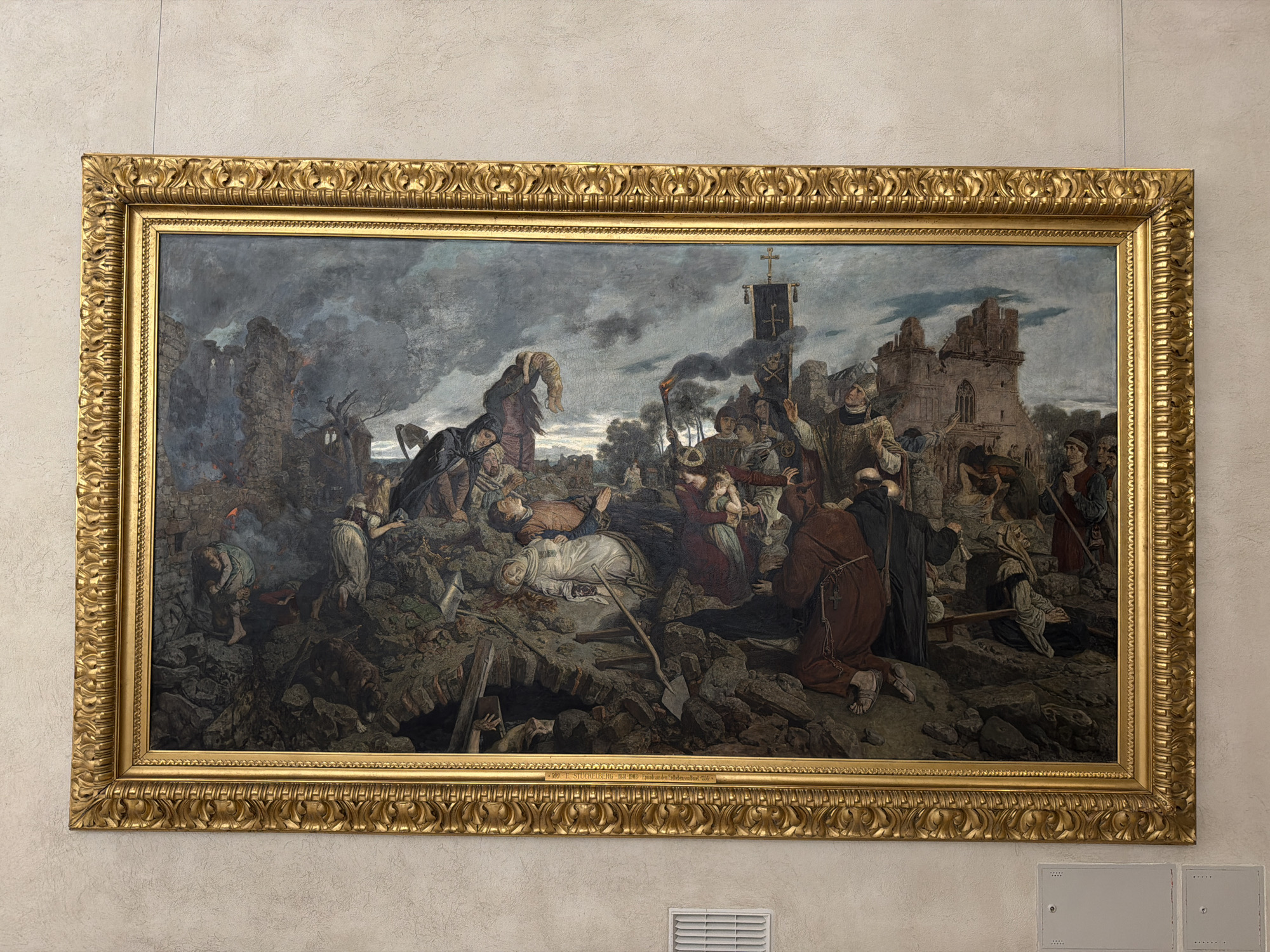 The image shows a large, ornate gold-framed oil painting depicting a chaotic and grim scene of destruction and death. The painting is hung on a textured light-colored wall. The subject matter appears to be a historical or religious event, with numerous figures in period clothing. In the foreground, there are many dead and wounded individuals lying among rubble and debris, suggesting a battle or disaster. Several figures are dressed in religious habits, including monks. Some individuals are actively engaged in rescue or aid, while others appear to be mourning or in distress.
In the background, ruined structures, possibly a church or castle, are visible against a dark, stormy sky. There are torches held by some figures, casting dramatic light and shadows. A banner with a cross is prominently displayed. The overall impression is one of tragedy, suffering, and religious devotion amidst devastation.
The text at the bottom of the painting, visible in one of the crops, reads "1899 F. STUCKELBERG 1831-1907 'Der Todten von Basel'". This identifies the artist as F. Stuckelberg and the painting's subject as "The Dead of Basel". Franz Stuckelberg (1831–1907) was a Swiss painter. This painting is indeed located in Basel, Switzerland, and it is housed in the Kunstmuseum Basel. The painting is titled "Der Tod der Bürgermeister von Basel" (The Death of the Mayors of Basel), also known as "Die Pest" (The Plague) or "Die Toten von Basel" (The Dead of Basel). It depicts the scene after the devastating plague in Basel in 1349.