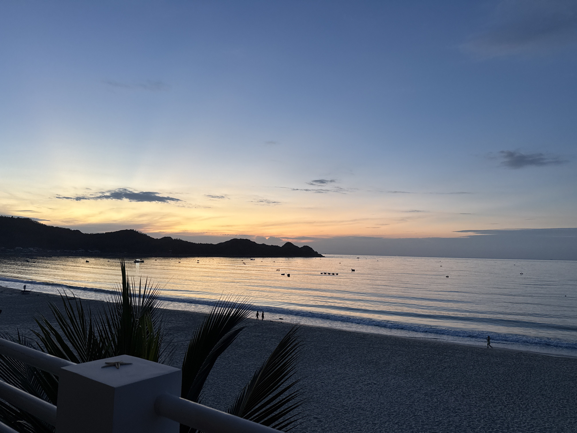 The image shows a beach scene at dusk. The sky is a gradient of blue and orange, with some clouds. The ocean is calm, with gentle waves. There are several boats on the water. The beach is sandy, with some palm trees in the foreground. There are a few people walking on the beach. The scene is peaceful and serene.