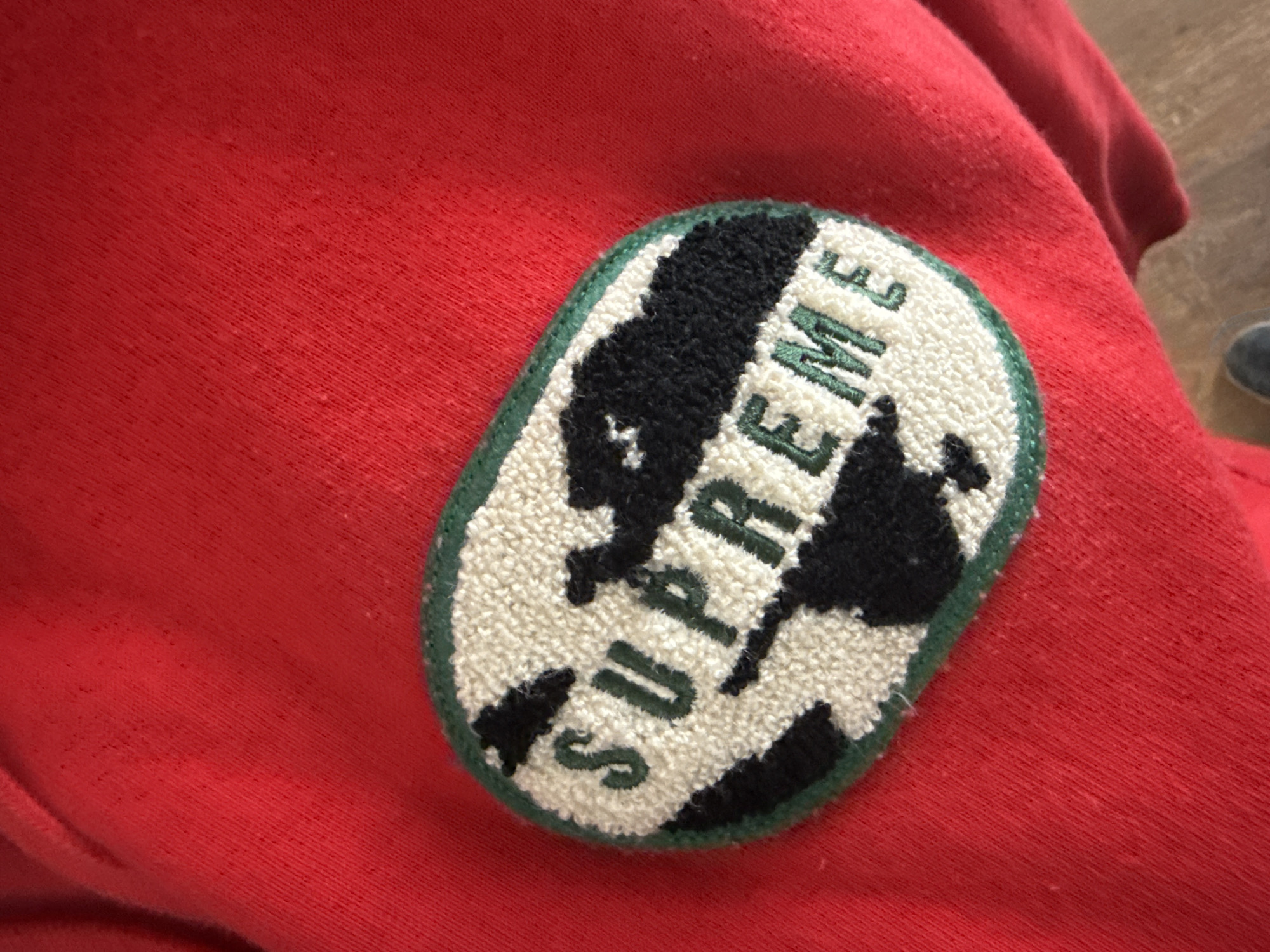 A close-up shows a red sweatshirt with an oval patch. The patch has a green border and features the word "SUPREME" in white, with black blocks obscuring parts of the text. The sweatshirt is wrinkled. The photo does not provide enough information to determine a location in Sofia, Bulgaria.