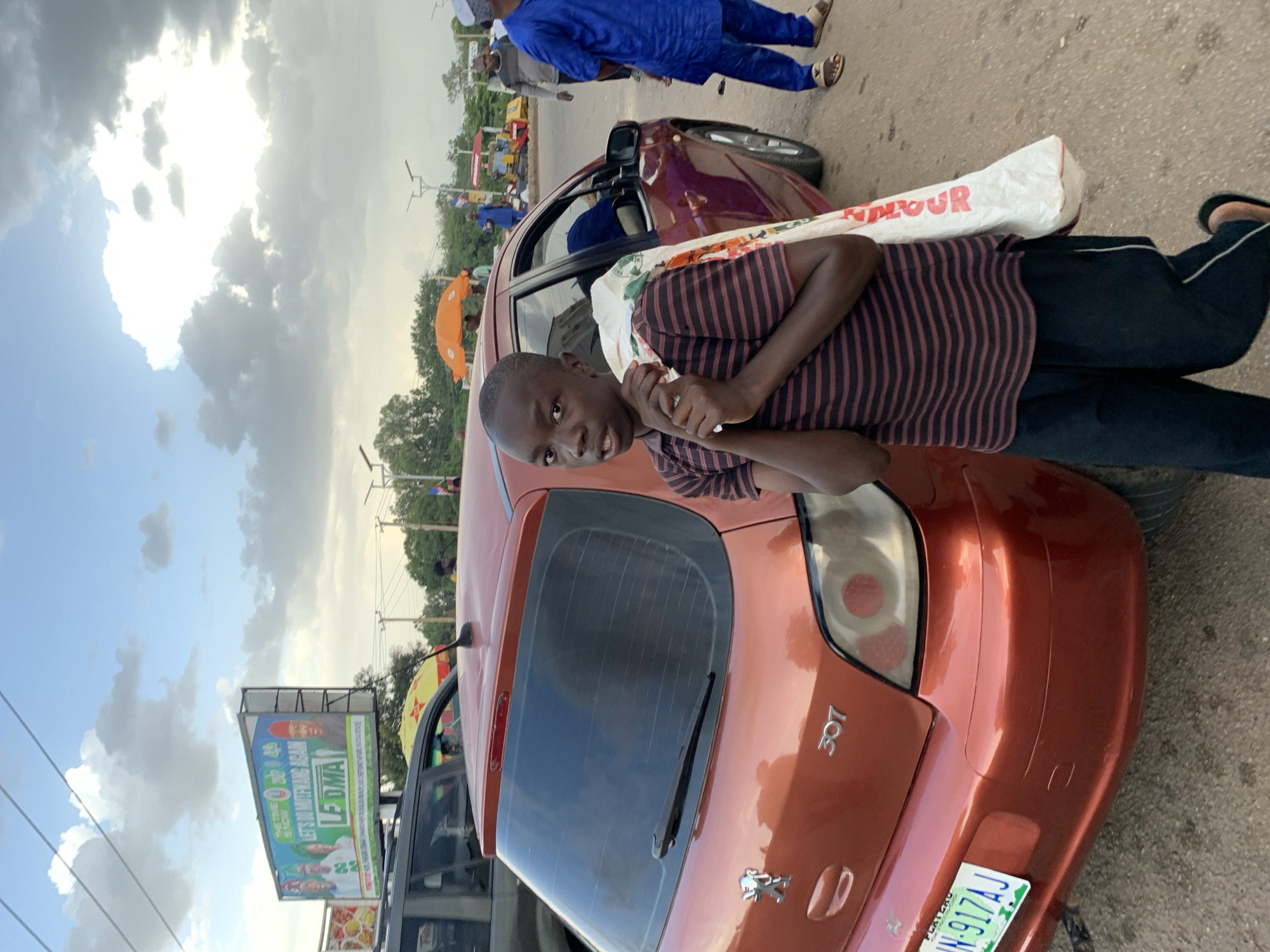The image shows a young boy standing next to an orange Peugeot 307 car. He is wearing a striped t-shirt and black pants, and he is carrying a white sack over his shoulder. In the background, there is a billboard with political imagery, traffic, and other pedestrians. The environment appears to be a street in the city of Jos, Nigeria. The sky is cloudy, suggesting it might be late afternoon or early evening. The billboard prominently displays "LET'S DO MUTFWANG AGAIN" and "LF DMA," indicating a political campaign or event.