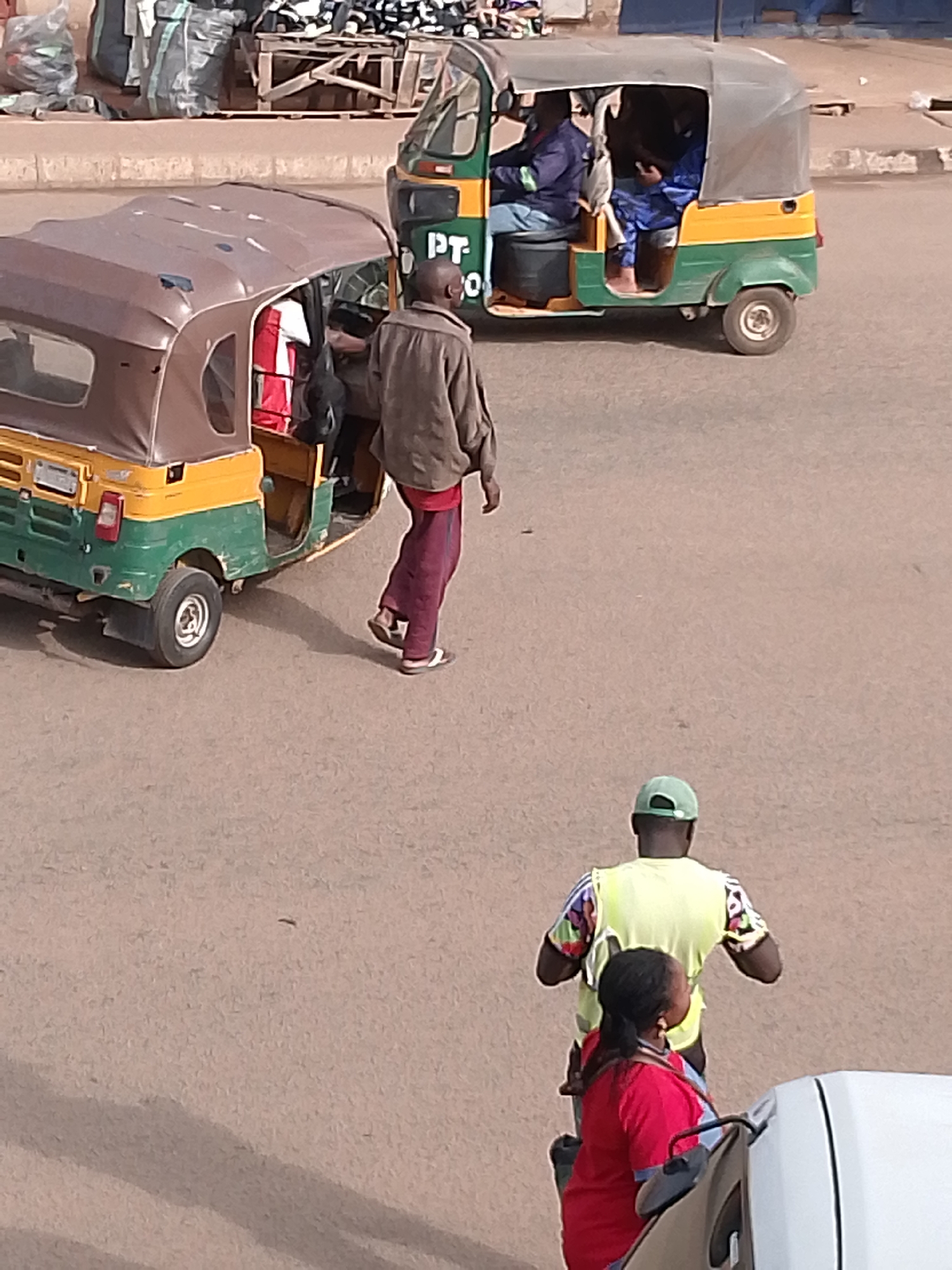 In Bukuru, Nigeria, a scene unfolds on a paved surface, likely a road. Two auto-rickshaws, commonly known as Keke Marwas, are present. The first, a yellow and green vehicle, is parked, and a man in a brown jacket and burgundy pants is walking away from it. The second Keke Marwa, also yellow and green, is in motion with two occupants visible inside. In the foreground, a person wearing a red shirt and a man in a bright yellow vest are positioned. The environment appears to be an urban setting with some bags and crates in the background, suggesting a marketplace or transport hub.