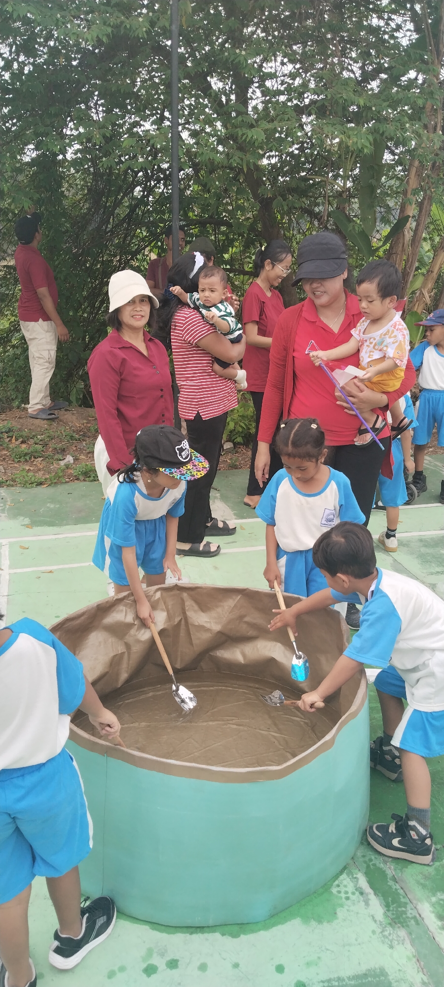 In Taban, Indonesia, a group of children and adults are gathered outdoors. The children, dressed in blue and white uniforms, are engaged in an activity at a large, light blue cylindrical tub filled with a light brown substance, possibly sand or mud, and water. They are using small shovels with wooden handles to scoop or mix the contents of the tub.
Several adults are present, some holding babies. One woman wears a red long-sleeved shirt and a black cap. Another woman wears a red shirt and a white bucket hat. A man in a red shirt and light-colored pants stands in the background. The setting appears to be a paved area with a green surface, likely a playground or outdoor recreational space, with lush green trees and vegetation in the background. The overall atmosphere suggests a children's activity or event.