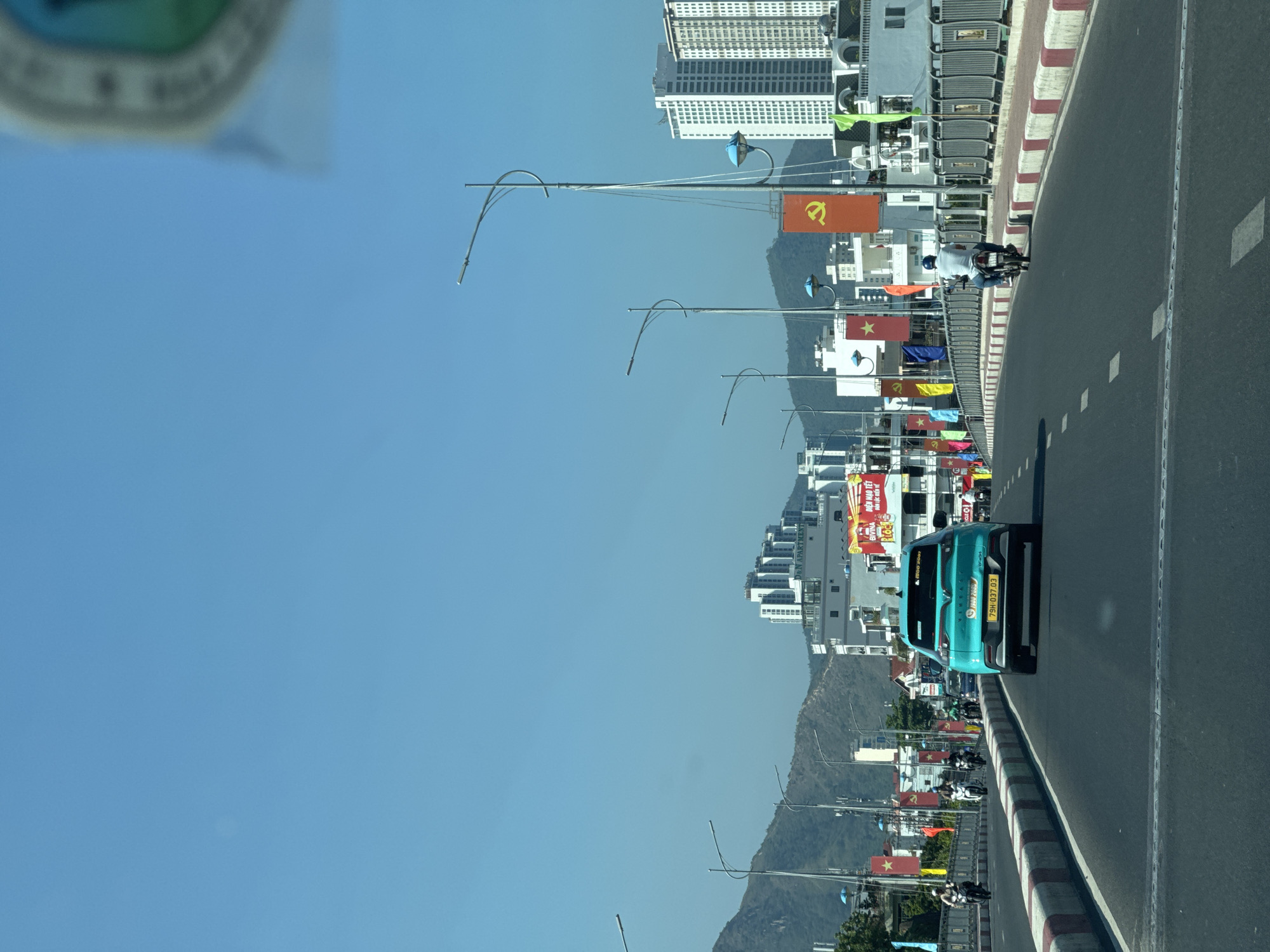 A turquoise car is driving on a road next to a sidewalk lined with streetlights and flags in an urban area. Buildings and mountains are visible in the background. The sky is clear and blue. A motorcycle is also on the road. The car's license plate reads "79H-037.03".