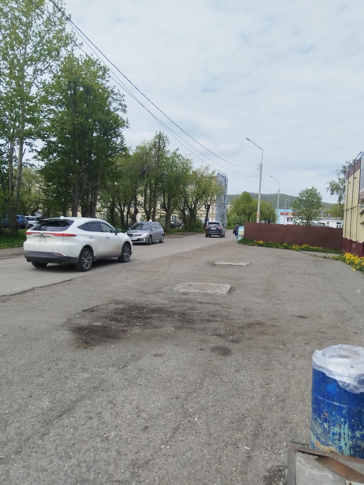 A wide shot shows a road in Vilyuchinsk, Russia, with a white SUV in the foreground. The SUV is parked on the right side of the road, facing left. It is a clean, modern vehicle with tinted windows. In the background, a few other cars are visible, as well as trees and buildings. The sky is overcast. The road is paved, with some potholes and cracks. There is a blue barrel on the right side of the image, covered with a plastic sheet. The overall impression is of a typical urban street scene.