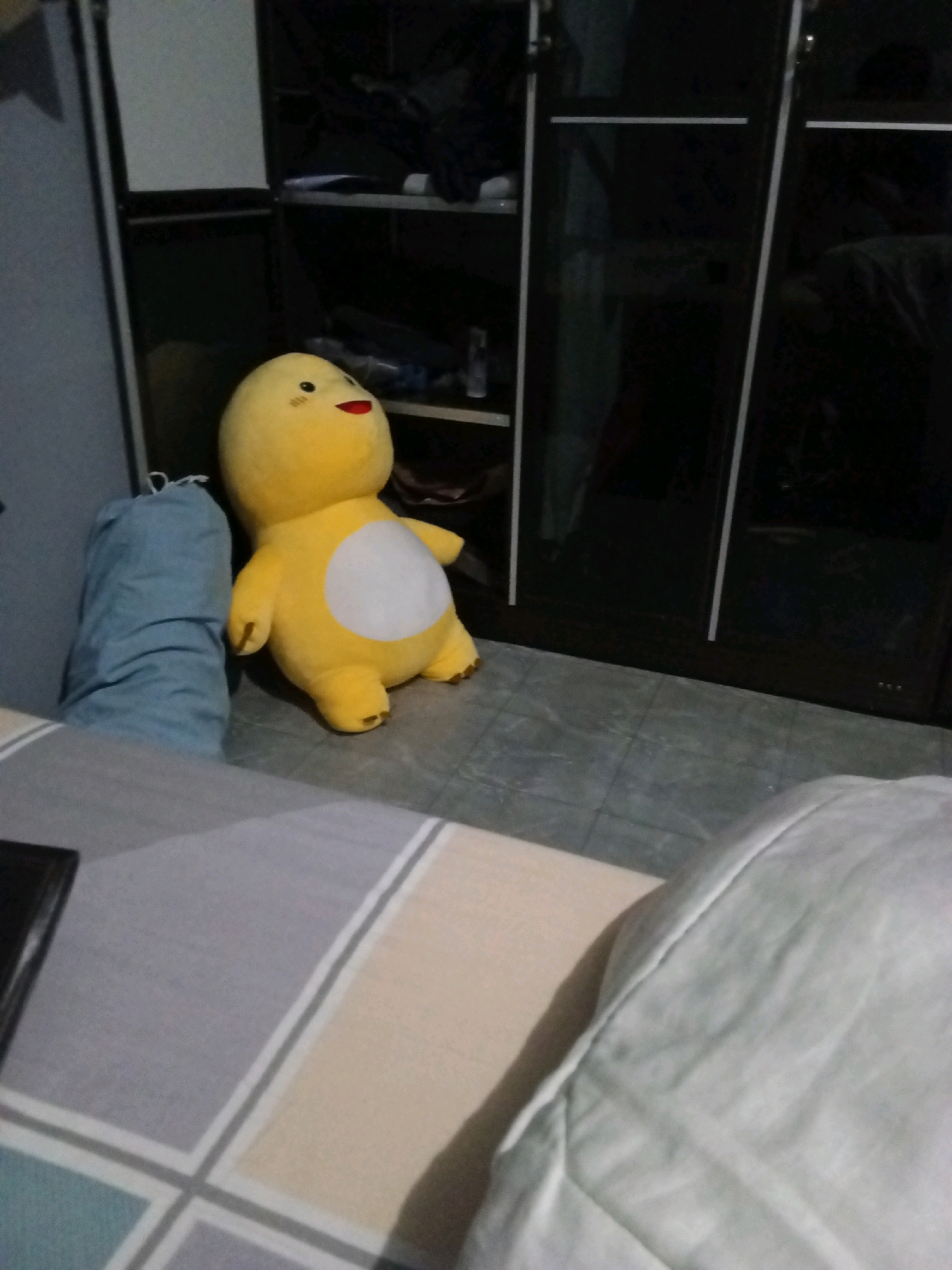 A yellow plush toy with a white circular belly, black eyes, red mouth, and pink blush marks on its cheeks sits on a gray tiled floor in front of a dark brown closet. To the left of the toy is a tall, light blue laundry bag. The foreground shows a bed with gray and beige striped bedding. The scene is dimly lit, suggesting an indoor environment. The location is identified as Gas Alam Badak I, Indonesia.