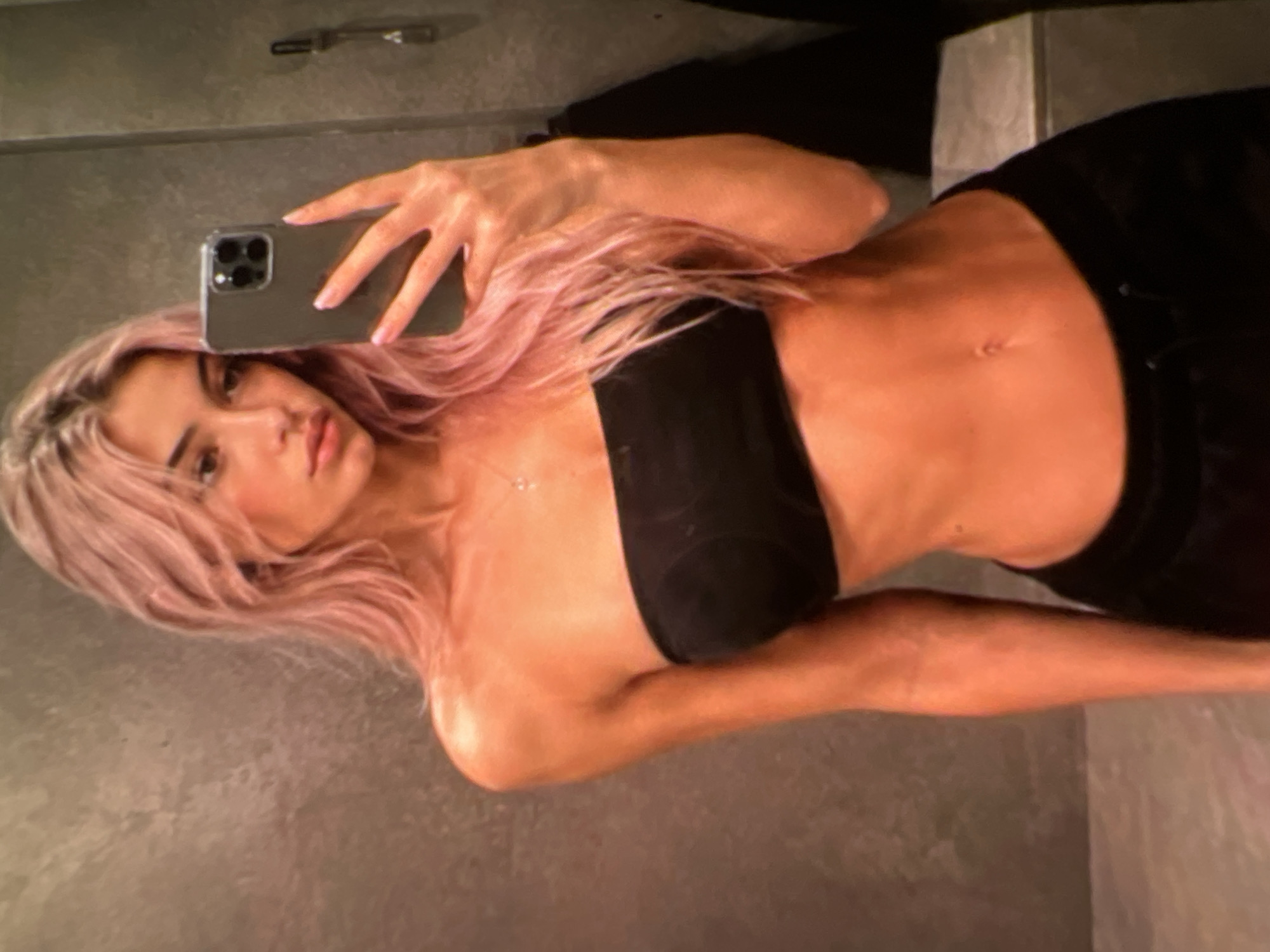 A young woman with long, wavy pink hair is taking a mirror selfie. She is wearing a black strapless top and black sweatpants. She is standing in what appears to be a bathroom, with a textured gray wall behind her. The lighting is warm and casts a glow on her skin. She is holding a smartphone in her right hand, angled to capture her reflection. Her left arm is resting by her side. She is looking at her phone's screen, with a neutral expression.