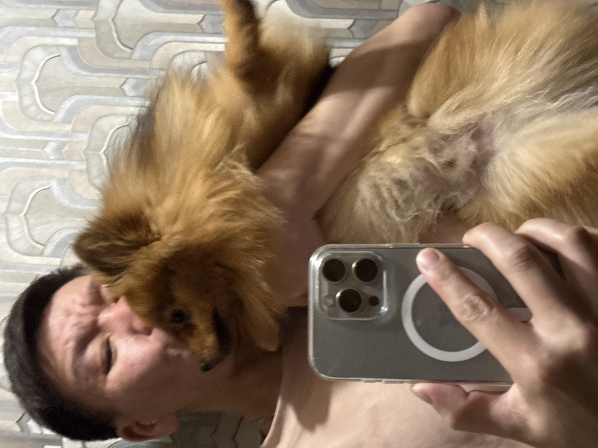 A selfie captures a person lying on a patterned surface with a brown, fluffy dog positioned over them. The person is holding a smartphone with a gray case, showing the camera lenses and a circular magnetic ring. The dog is close to the person's face, possibly interacting. The backdrop is a wall or floor covering with an off-white and gray geometric pattern. The location is purportedly in the city of Khimki, Russia, but this cannot be verified from the image alone.