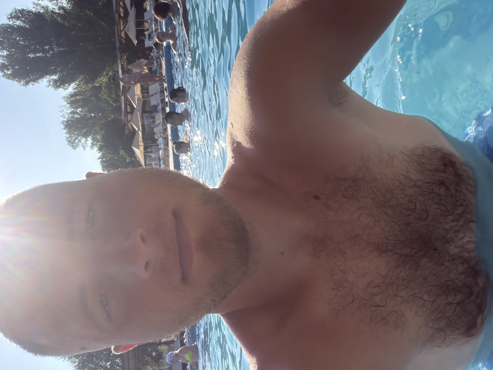 A man, appearing to be in his late twenties or early thirties, takes a selfie in a swimming pool. He is bald, with short stubble on his face and a visible patch of dark hair on his chest. The sunlight beams brightly from the upper left corner, creating a lens flare effect and partially obscuring his face. He is submerged in clear, blue water, with other people visible in the background, some swimming and others relaxing by the poolside. Umbrellas and lounge chairs are set up around the pool. The environment suggests a sunny day at an outdoor public pool, potentially in a resort or recreational area.