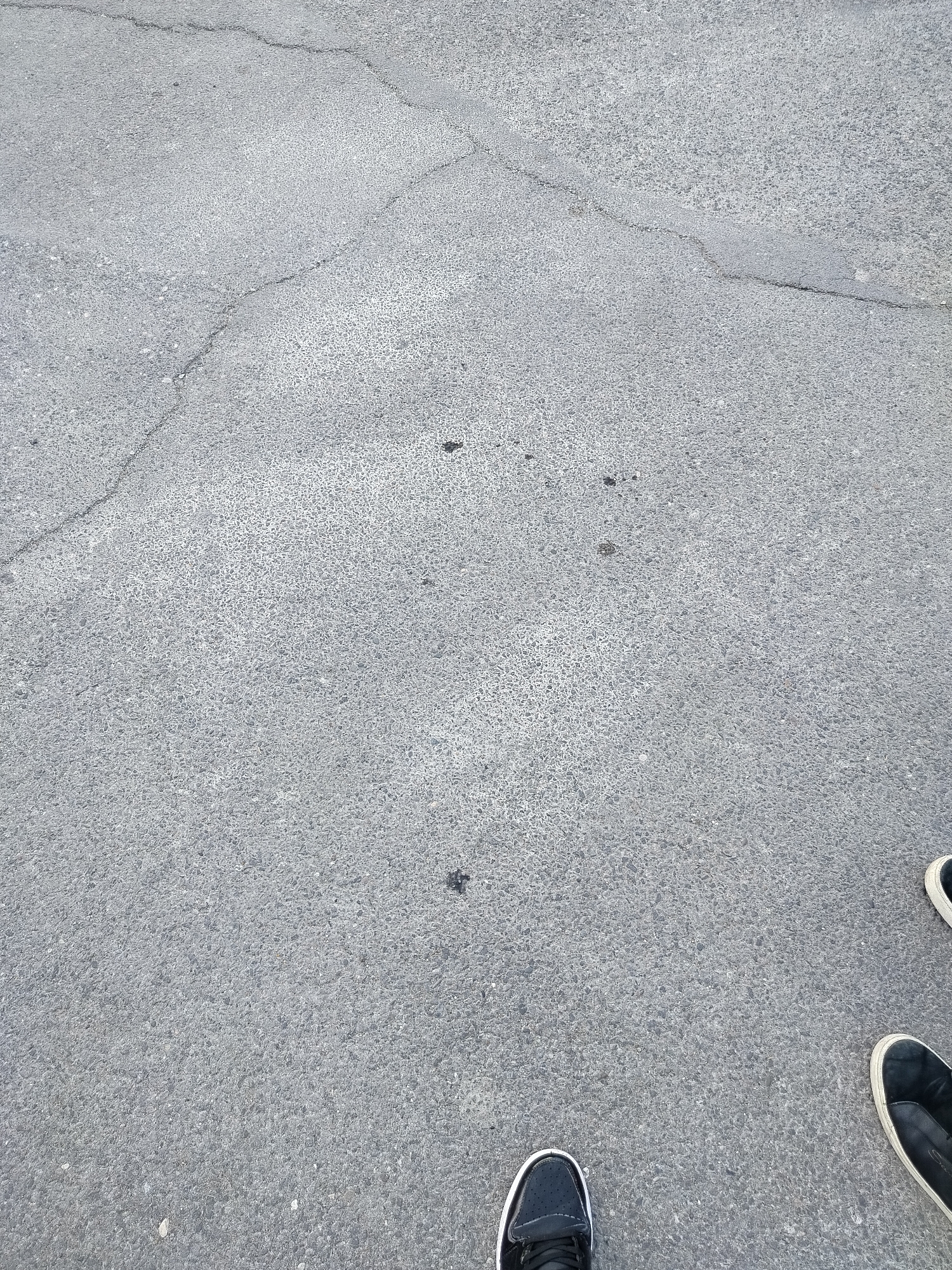 The image shows a section of paved asphalt ground with cracks. There are scattered dark spots on the pavement. Two pairs of shoes are visible in the lower part of the image. One pair of shoes are black with white trim and the other pair of shoes are black with white soles. The image provides no information regarding location in Yerevan or any subjects or actions. The environment is exterior, and the lighting appears to be natural.