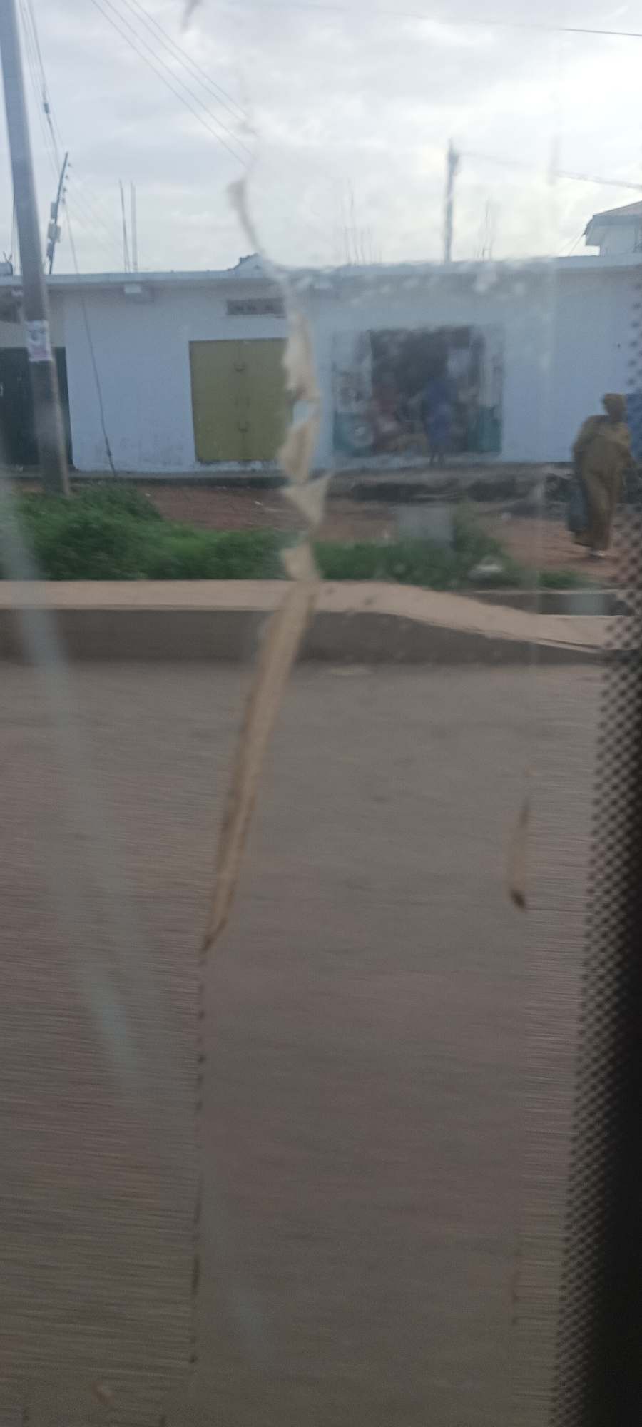 The image captures a street scene in Bukuru, Nigeria, viewed through a window marked with prominent streaks and reflections, suggesting it was taken from a vehicle, possibly in motion. The foreground consists of a blurred, dark grey road and a raised concrete curb. Beyond the curb, a strip of green grass and dirt is visible.
In the midground, a light-colored, single-story building dominates the scene. It features a yellowish-green double door with dark, curved handles on the left side of its facade. To the right of the door, an open storefront reveals a darker interior with blurry figures, possibly customers or vendors, and what might be a small, light-colored vehicle or tricycle partially visible. A person wearing a long, light brown garment is standing or walking on the far right, partially obscured.
The background shows an overcast sky with scattered clouds. Utility poles and power lines are visible on the left side, extending upwards. The overall lighting suggests it is daytime. No legible text is visible anywhere in the image.