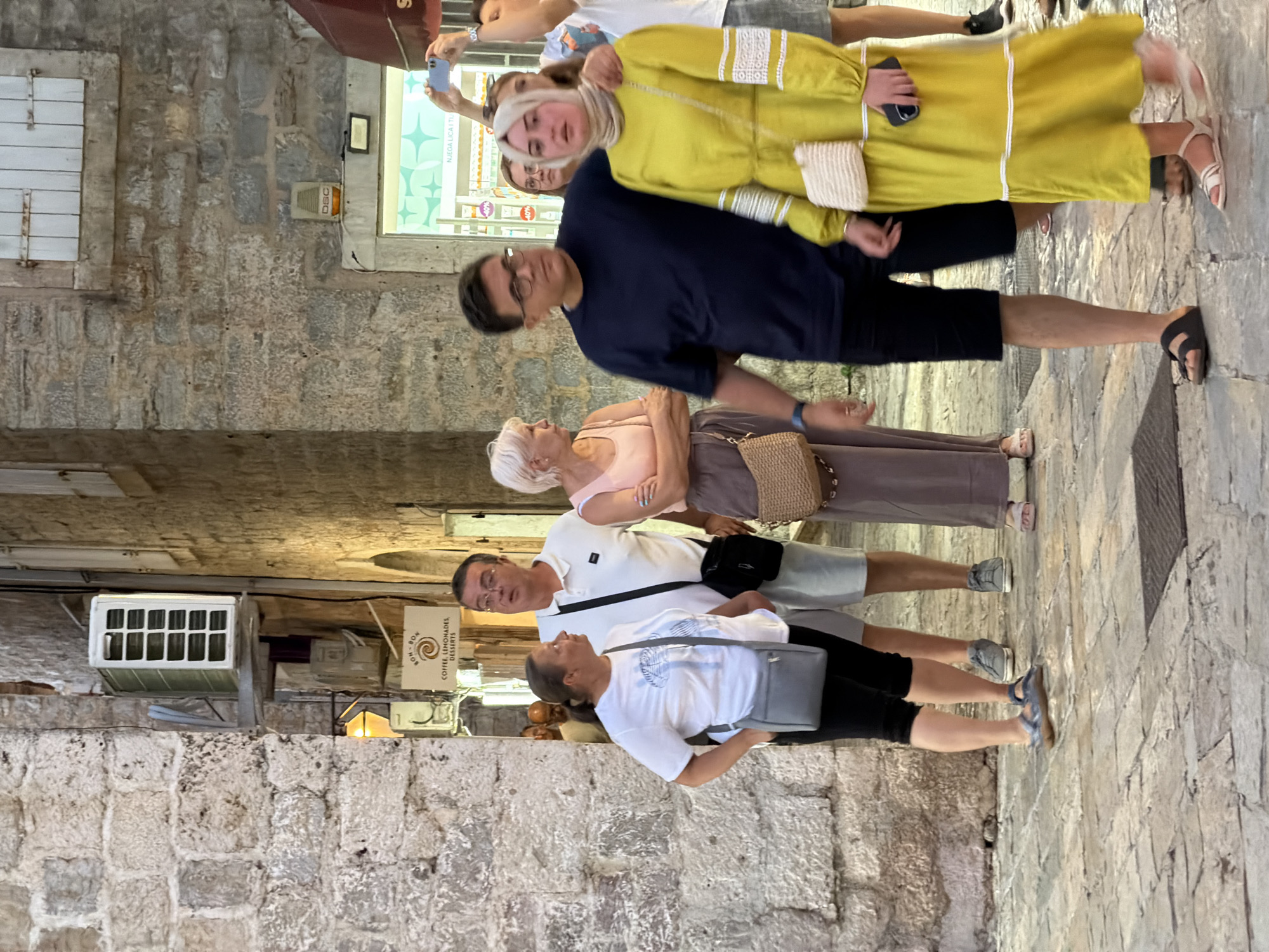 In Budva, Montenegro, a group of people are walking down a narrow, cobblestone street lined with stone buildings. The scene appears to be in the evening, with artificial lighting illuminating the area.
In the foreground, a woman in a yellow dress is walking towards the viewer, holding a smartphone. To her left, a man in a dark blue t-shirt and black shorts is walking with his arm around her. Behind them, a woman with blonde hair in a light pink tank top and a woman in a white t-shirt are visible. Further in the background, a man in a white polo shirt and shorts is also walking. Another man in a white t-shirt is seen in the distance.
The street features several air conditioning units mounted on the walls, and a sign that reads "BON BON COFFEE, LEMONADES, DESSERTS". The architecture suggests an old European city.