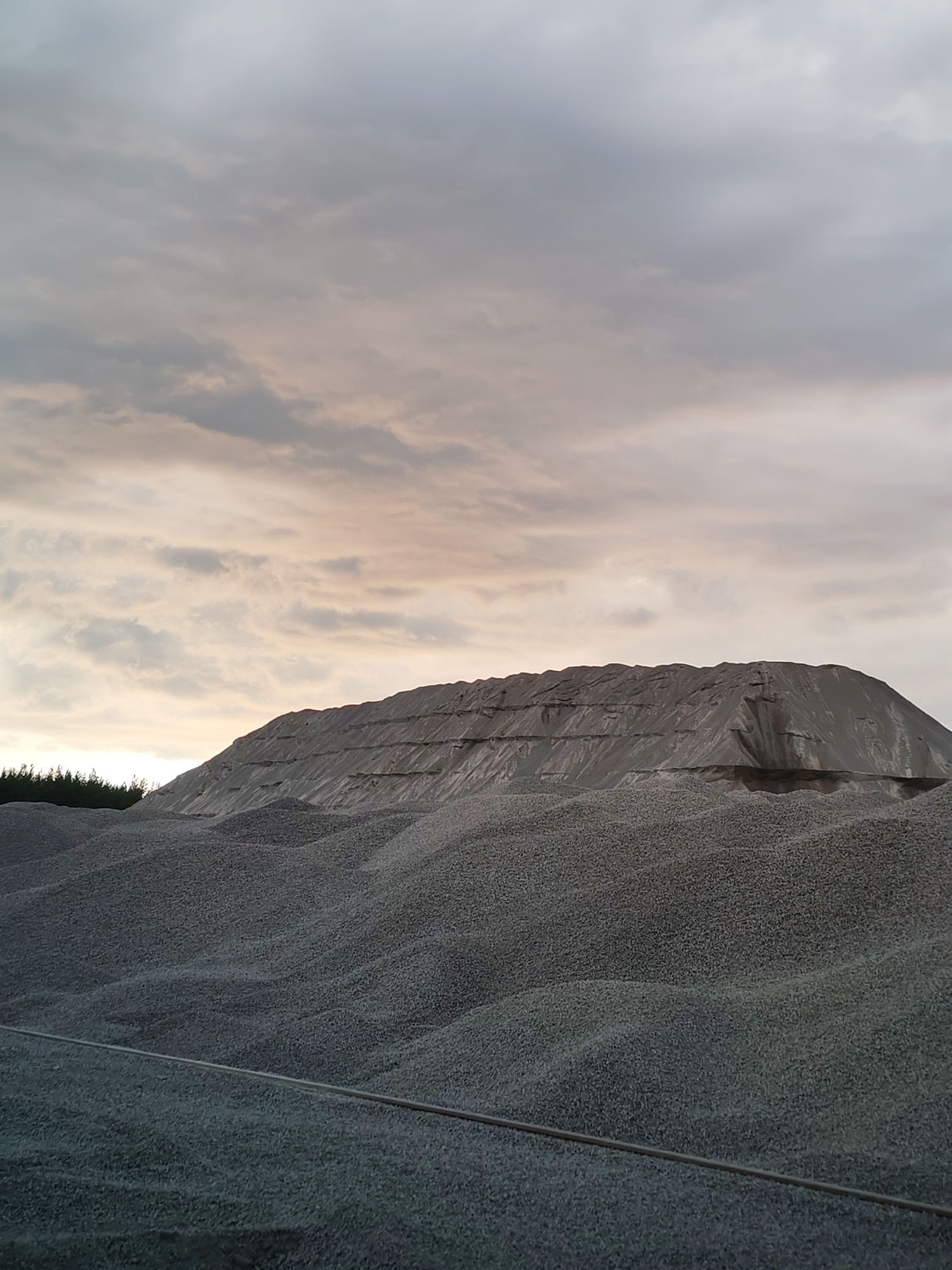 The image depicts a large pile of gravel in Shalsky, Russia, under a cloudy sky. The gravel mountain is covered in part by a grey tarp, creating a layered effect on its peak. In the foreground, more gravel is visible, with a metal wire or cable running horizontally across it. To the left, a dark line of trees suggests a forested area bordering the gravel deposit. The lighting indicates it may be dawn or dusk. There are no people or animals visible in the scene.