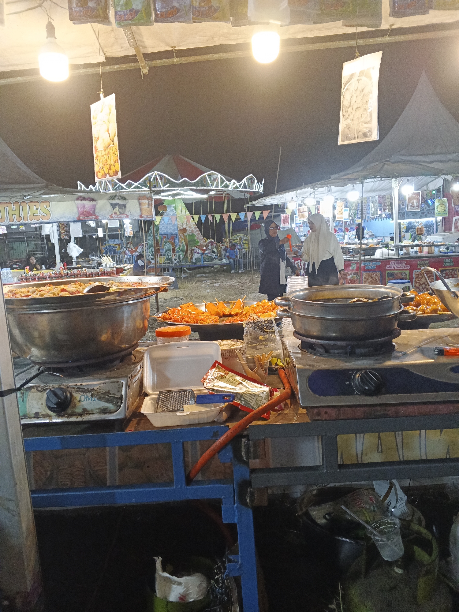 An outdoor food stall is depicted at night, likely a marketplace or fair in Sukabumi, Indonesia, given the presence of food vendors and various attractions in the background, including what appears to be a carousel or small amusement ride. The foreground features a food stall with two large metal cooking pots on gas stoves. One pot is filled with a reddish-brown, likely fried, food item, possibly sausages or similar snacks, and a large metal spoon is resting inside. To the right of this pot, another pot sits on a stove, containing dark liquid. In front of the stoves, there are various food items arranged, including skewers of orange-colored food, possibly fried chicken or meatballs, on platters. Styrofoam containers, a grater, and other kitchen utensils are also visible.
Two women are standing behind the food stall, one wearing a black jacket and sunglasses, and the other a white hijab. They appear to be interacting with each other, possibly as vendors or customers. In the background, more stalls and structures are visible, adorned with colorful banners and lights, suggesting a festive atmosphere. The overall scene is characteristic of a bustling night market or street food event.