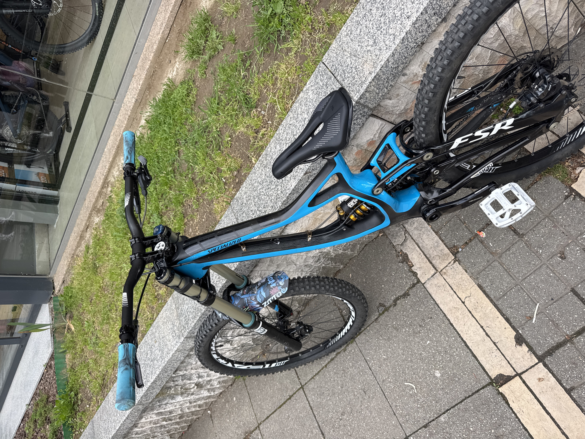 A blue and black Specialized mountain bike is prominently featured, parked outdoors and leaning against a grey granite curb. The bike is a full-suspension model, likely designed for downhill or enduro riding, evidenced by its robust frame, dual-crown (or heavy-duty single crown) front fork, and rear coil shock. The frame displays "SPECIALIZED" on the top tube and "FSR" on the rear swingarm, which also has "SICK!!" and partially visible "DESIRE" text. A Cane Creek Double Barrel ("DB") shock is visible at the rear.
The handlebars are equipped with distinctive blue patterned grips, and a small patterned mudguard, possibly branded "ZATTO", is attached to the front fork. The wheels feature knobby tires with white branding, possibly "ZUUM", on the rims. The drivetrain includes a "DESCENDANT" crankset and white/silver flat pedals. The black saddle has a unique white polka-dot pattern on its sides.
The bike is positioned on a concrete sidewalk composed of various sized slabs, with a strip of green grass visible beyond the curb. A large glass window to the left reflects parts of another bicycle, hinting at a nearby bike shop or store. The scene is illuminated by natural light, suggesting daytime, possibly under an overcast sky as there are no harsh shadows. Minor litter is scattered on the sidewalk. The setting is an urban or suburban environment in Blagoevgrad, Bulgaria.