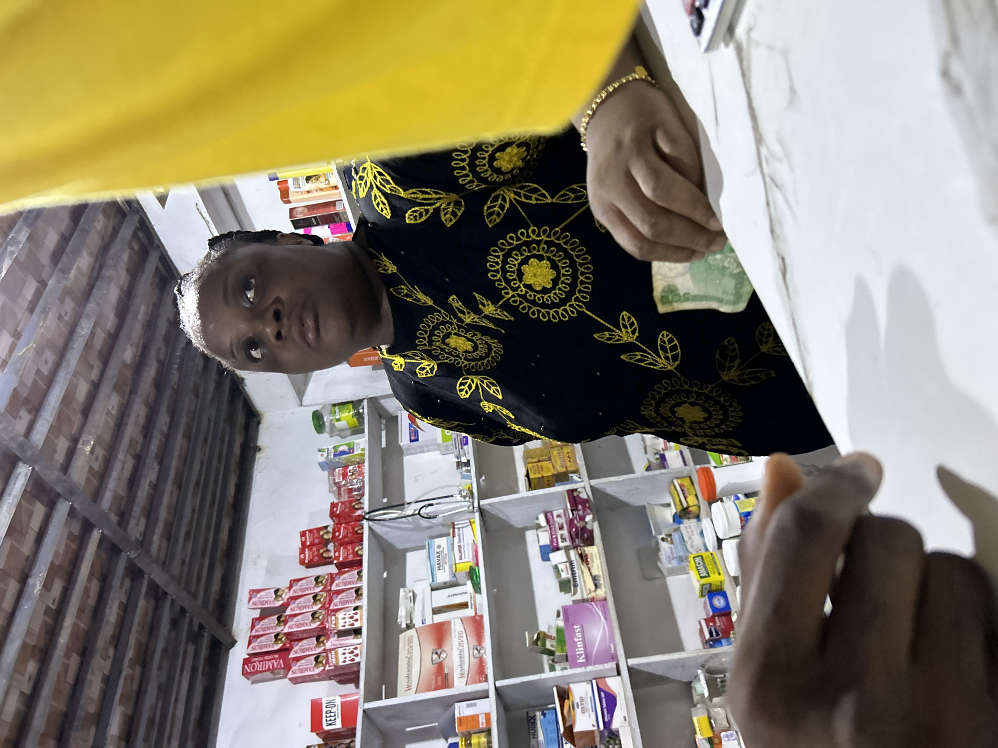 The image depicts the interior of a pharmacy in Ihiagwa, Nigeria. A woman in a black and yellow patterned shirt stands behind a counter, holding a green bill. The shelves behind her are stocked with various medications, including boxes of VAMIRON, NUDEX, Klinfast, Havax and Emtrim as well as other pharmaceutical products. The ceiling of the pharmacy has an exposed metal frame. Another person's hand appears in the foreground, possibly a customer. The scene suggests a transaction taking place.